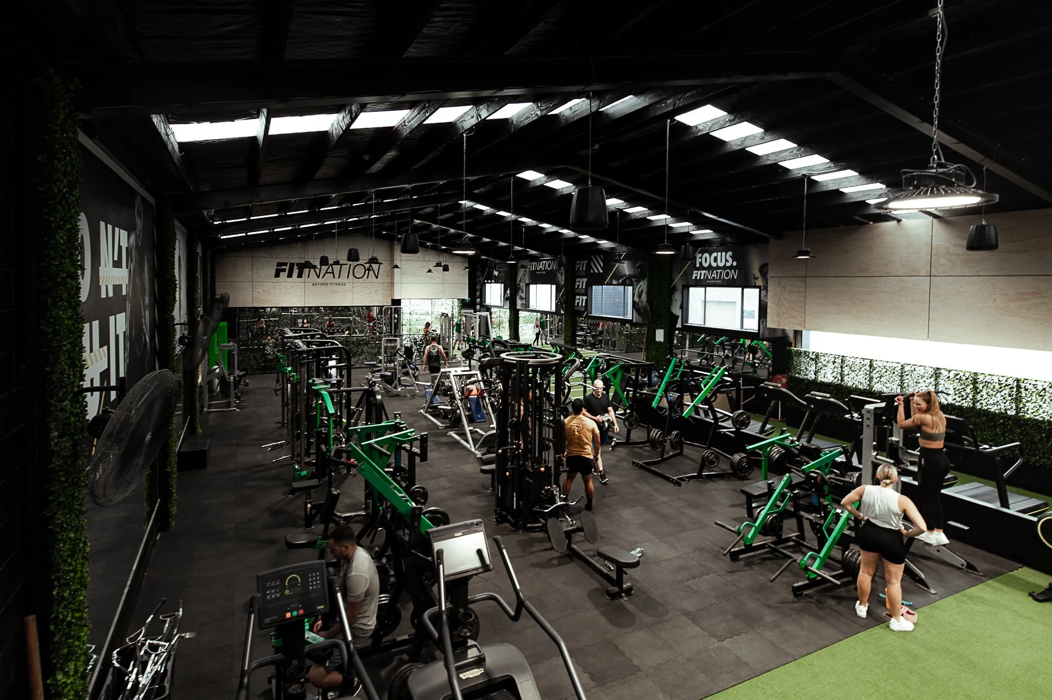 Inside a modern gym with various fitness equipment, including treadmills, weight machines, and benches. Several people are working out or stretching, and the gym has black walls with motivational signs like 'FITNATION' and 'FOCUS.'
