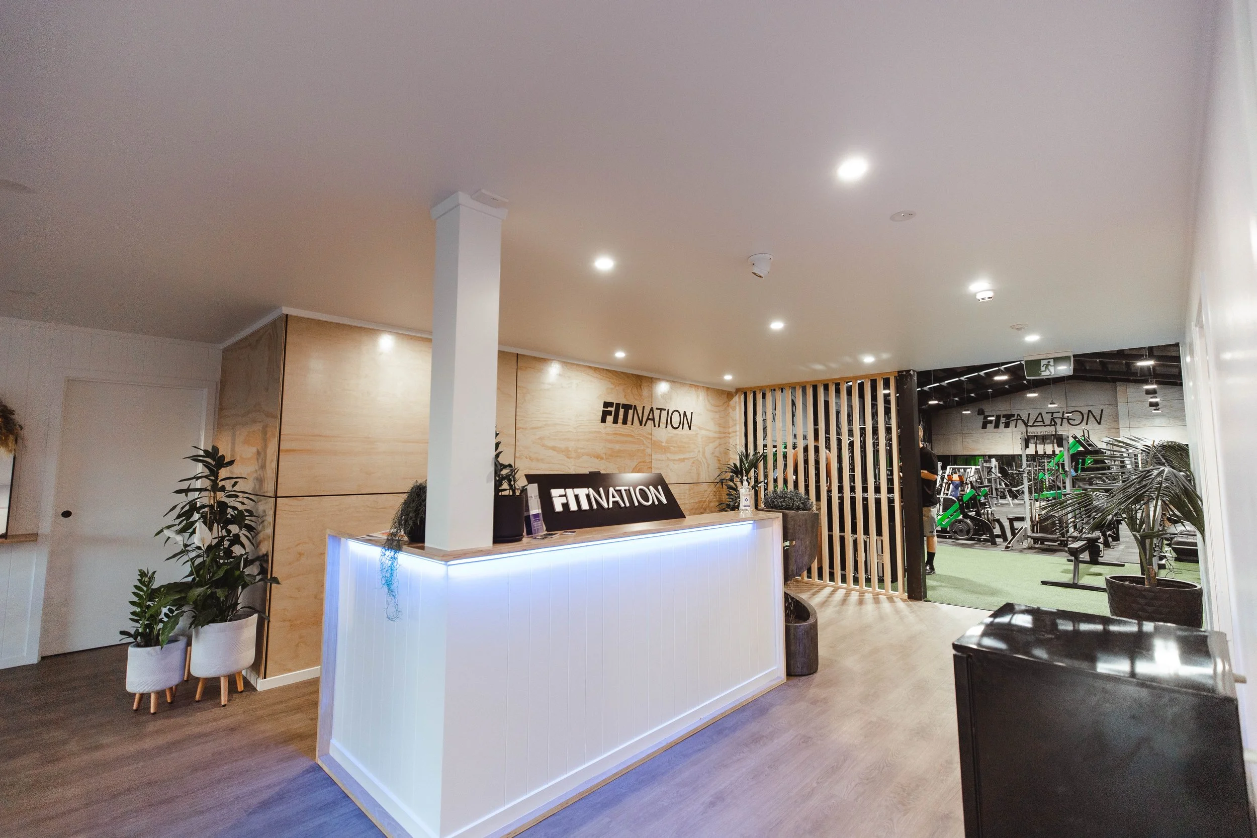 Reception area of a fitness center with a wooden wall, potted plants, and a sign that reads 'FITNATION'; gym equipment visible through a glass partition in the background.
