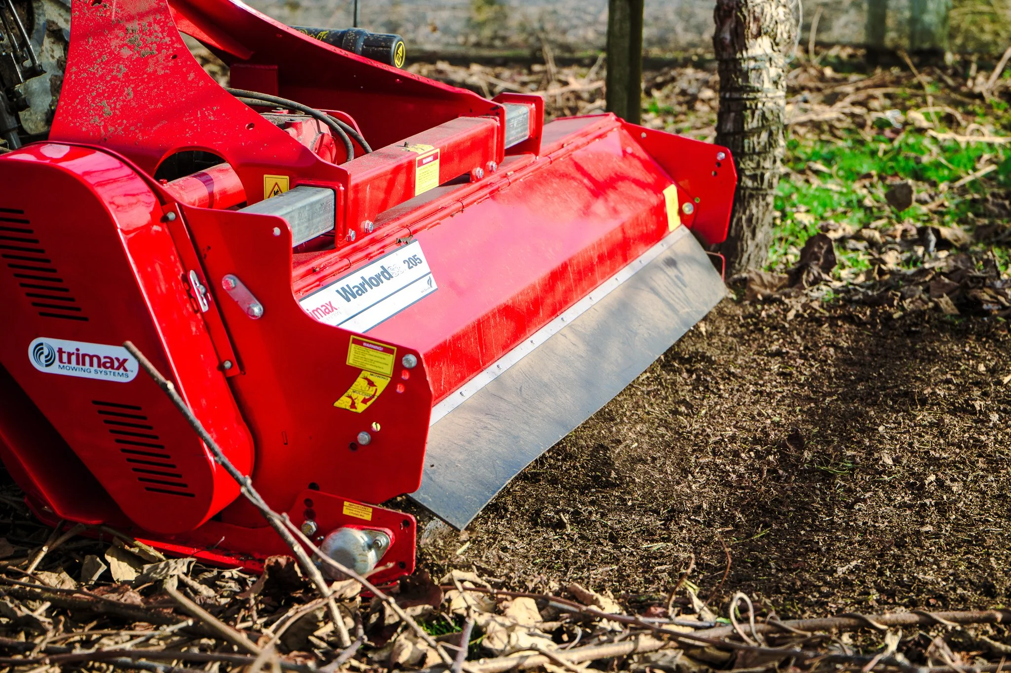 A red wood mulcher with a metal blade in an outdoor setting, surrounded by soil, leaves, and a tree trunk.