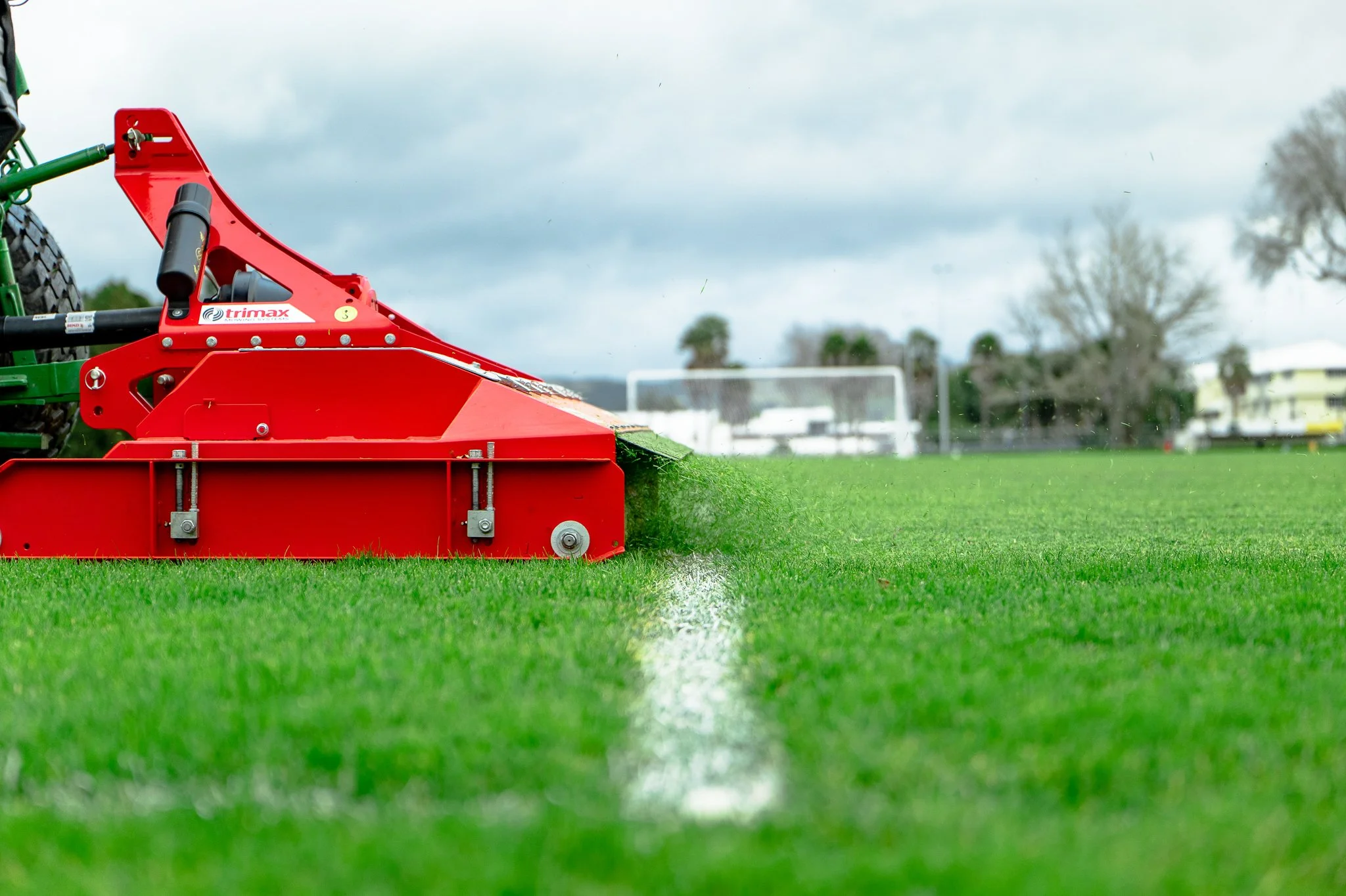 A red and green field grooming machine is cutting grass on a sports field, with goalposts and trees in the distance under a cloudy sky.
