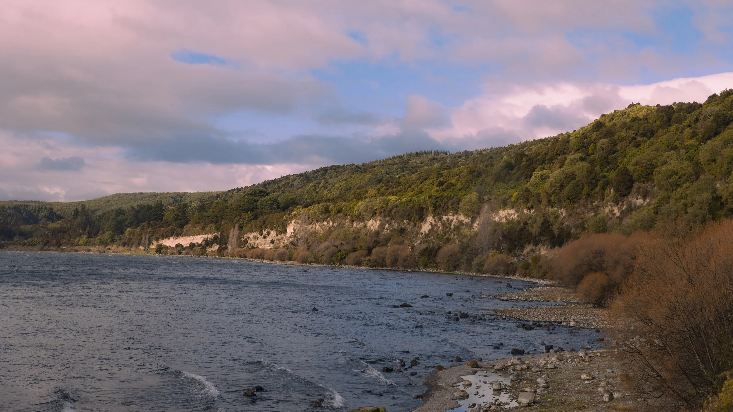 View of a river with gentle waves, rocky shoreline, and forested hillside under a partly cloudy sky.