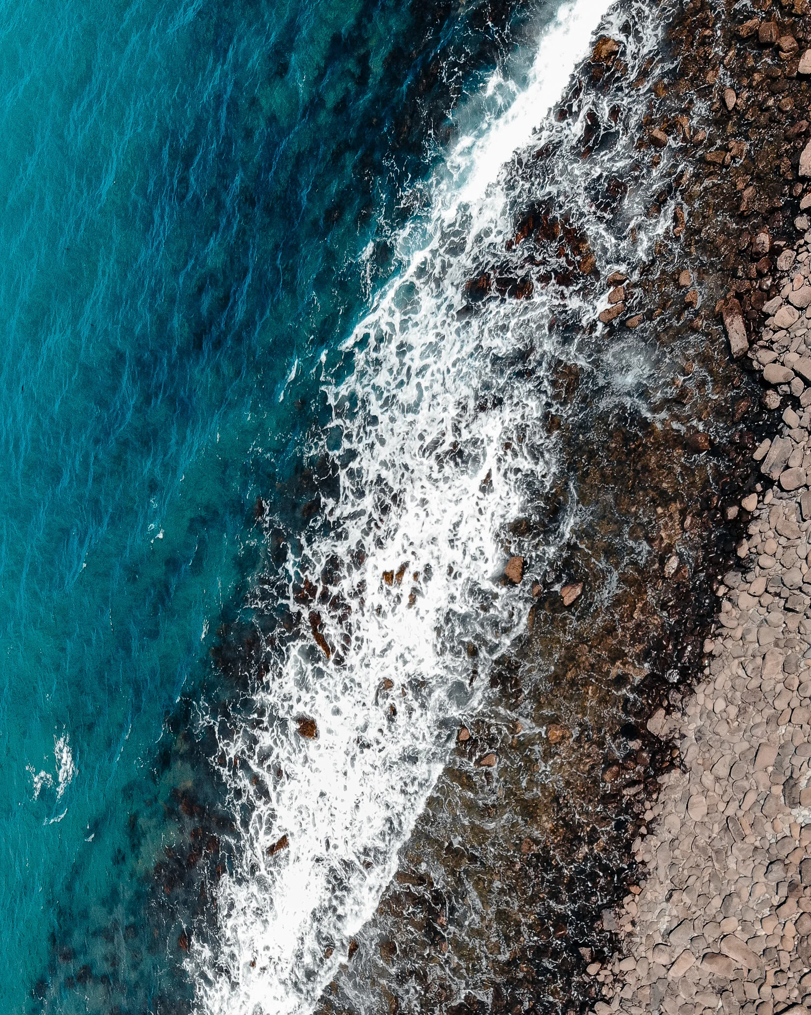 An aerial view of the shoreline showing turquoise ocean water meeting dark rocky and sandy beach.