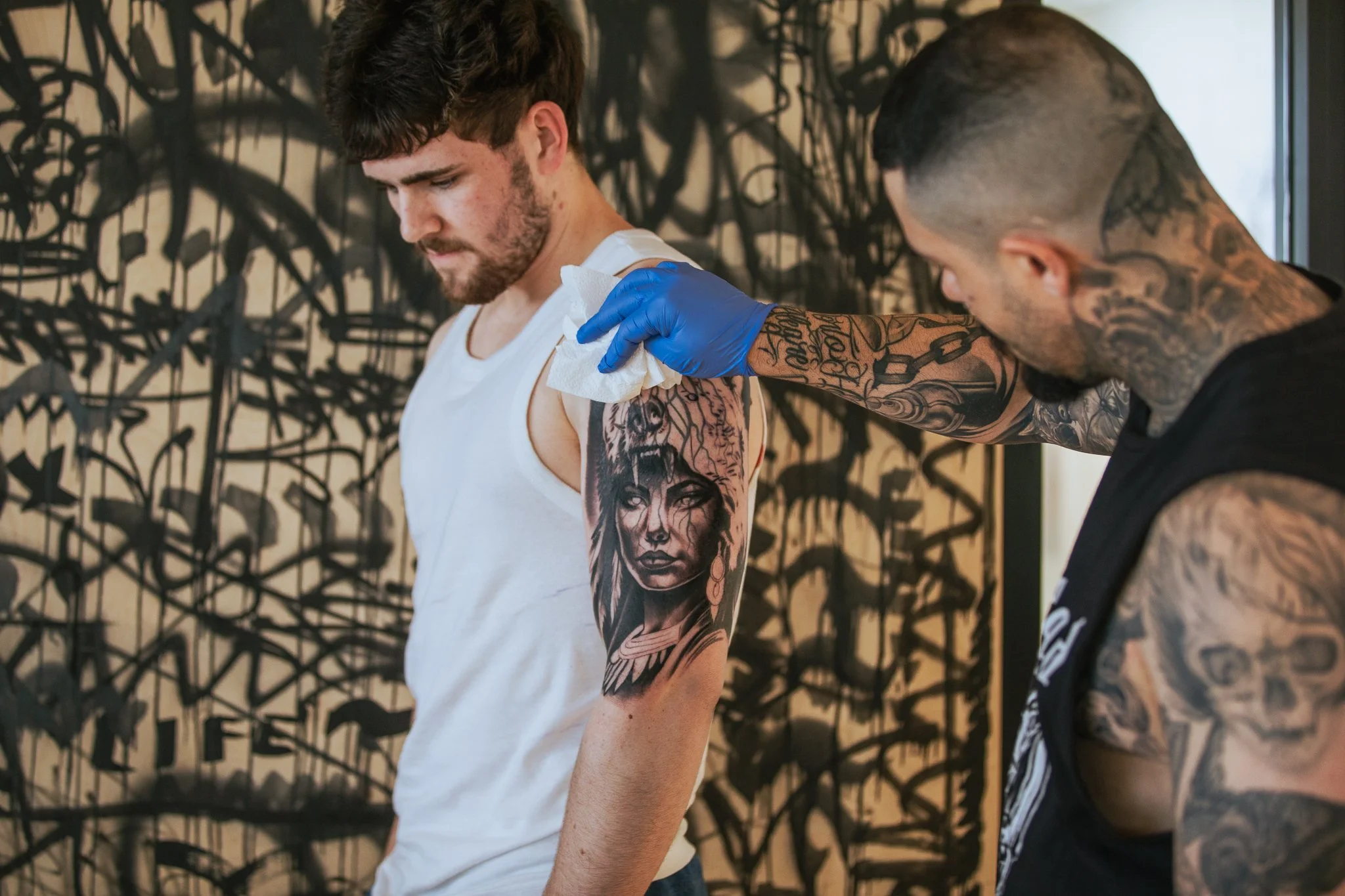 A man with tattoos receiving a shoulder tattoo from a tattoo artist in a tattoo studio.