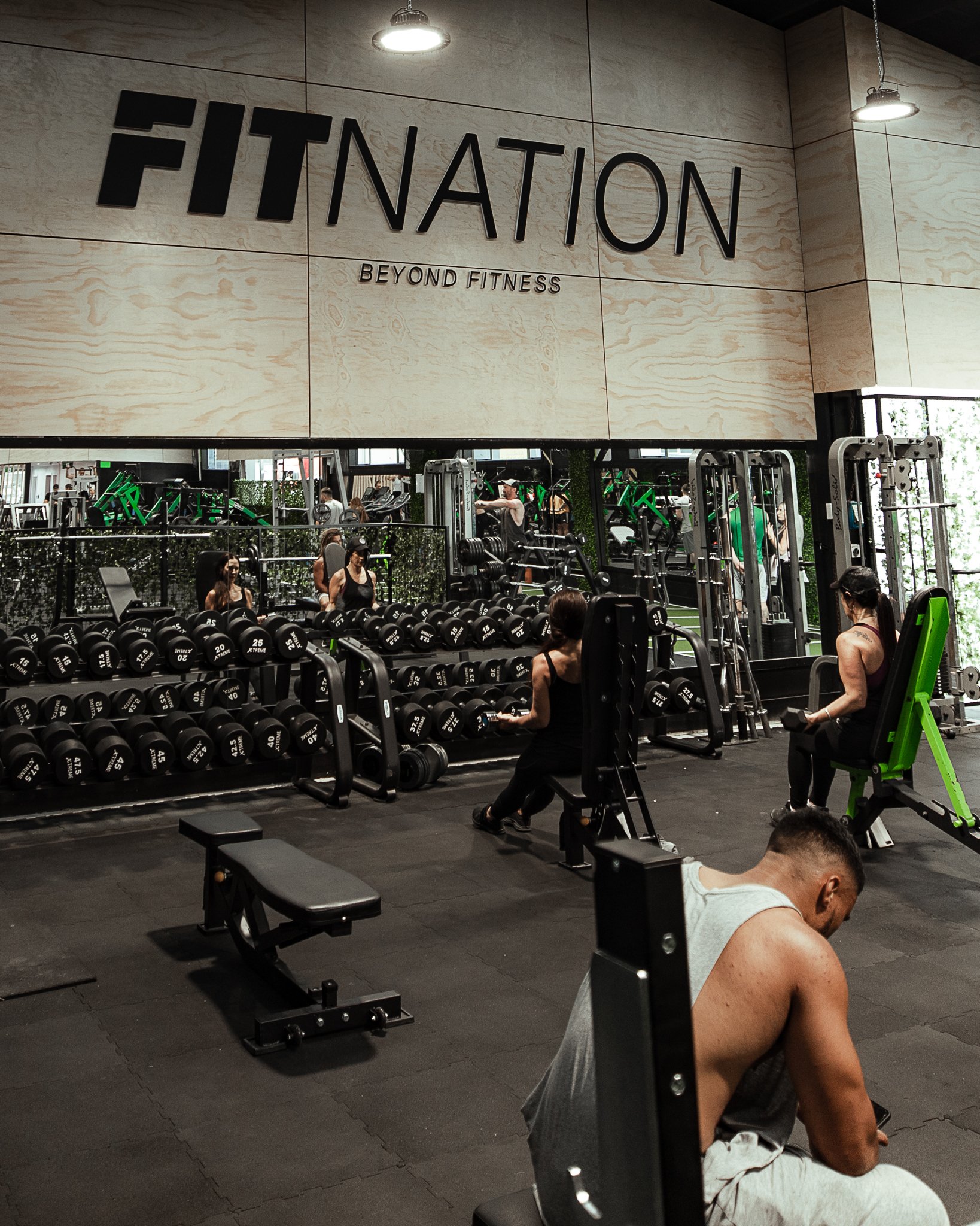 Inside a gym with people working out on equipment, a large sign above reads 'FITNATION BEYOND FITNESS'.