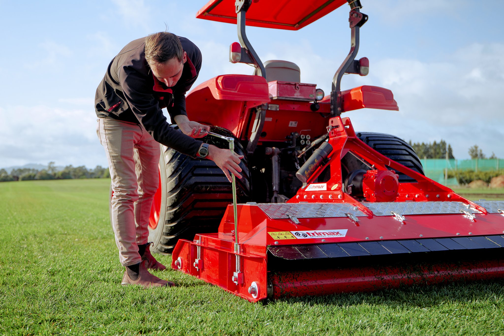 A man inspecting a large red agricultural machine on a green field with trees and cloudy sky in the background.