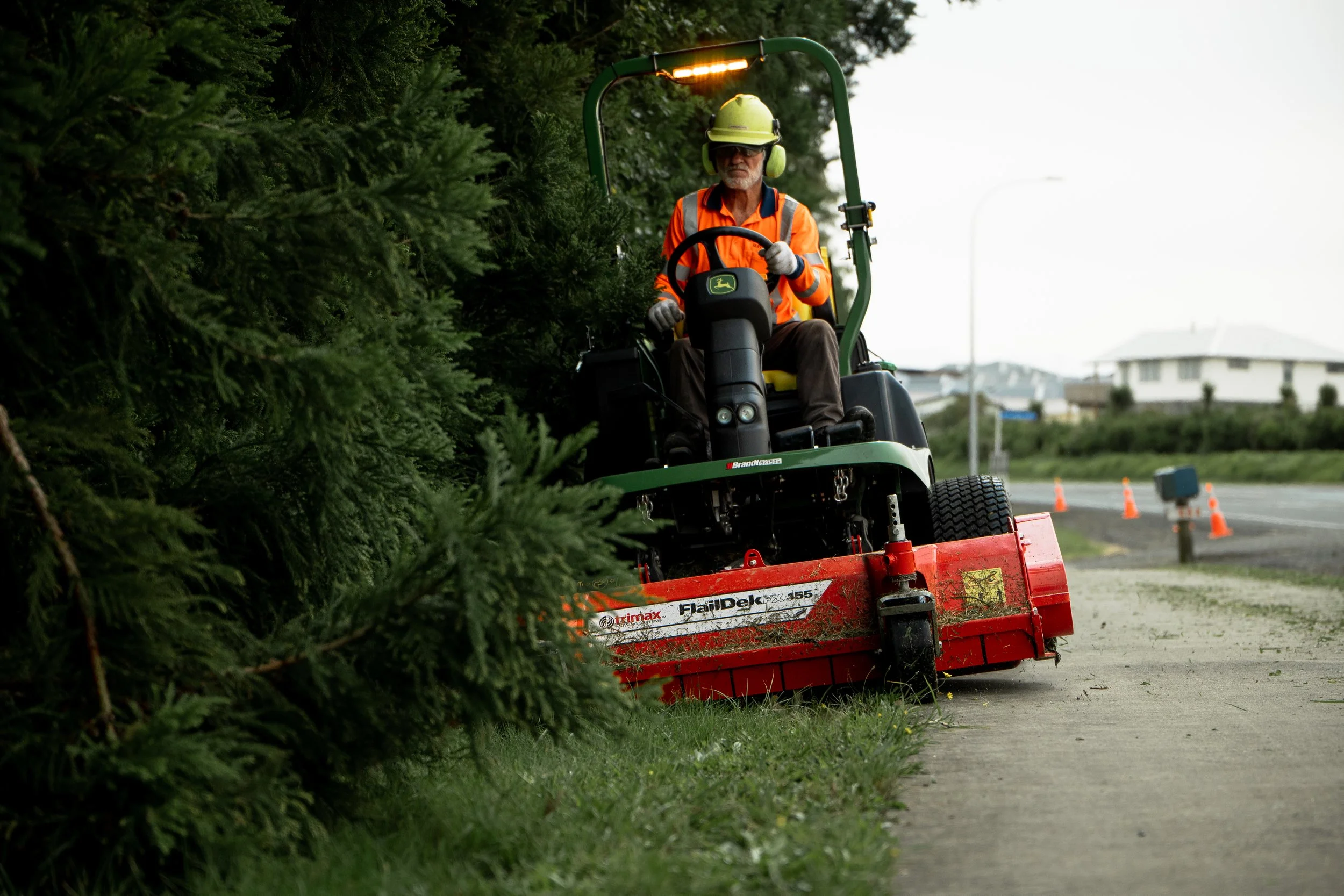 A man operating a John Deere lawn mower trimming grass near a sidewalk with trees on the side, wearing a yellow helmet, orange safety vest, and ear protection. There are orange cones in the background on an overcast day.