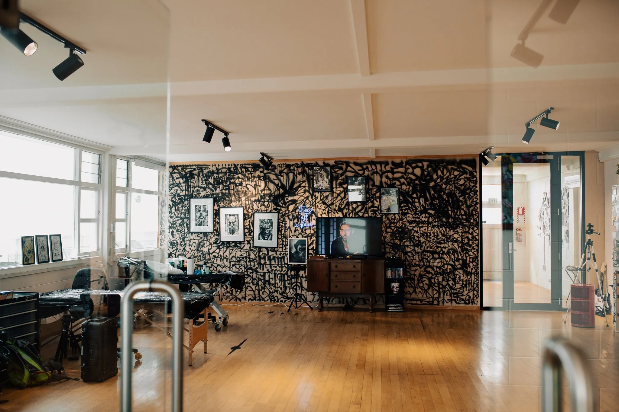 An indoor art studio or gallery space with a patterned black and brown wall, framed photographs, and a television showing a man. The floor is wooden, and there are windows with natural light, with art supplies and equipment visible.