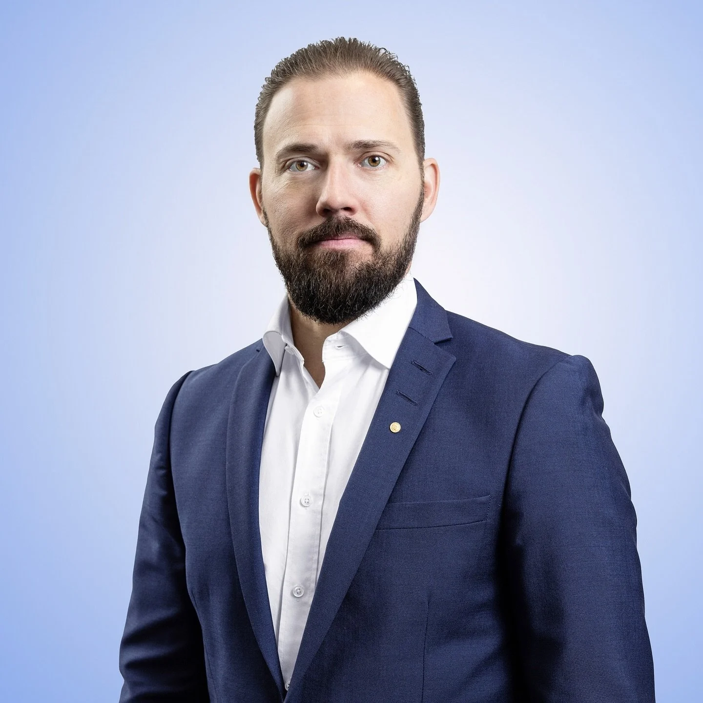 Henkilö muotokuvassa, joka katsoo kameraan. Tausta vaaleansininen. Hänellä on parta ja hän on pukeutunut valkoiseen paitaan ja tumman siniseen bleiseriin.