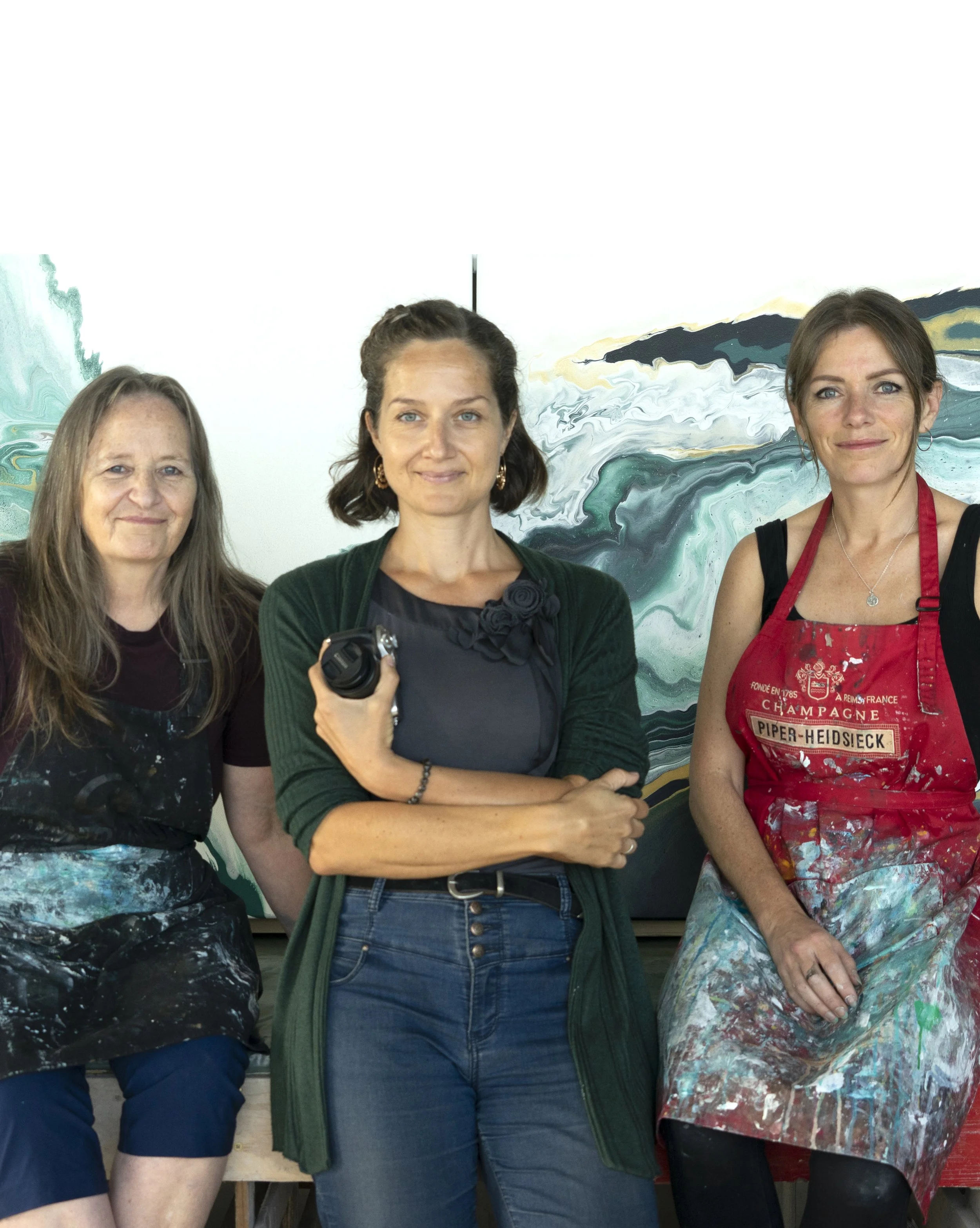 Three women standing in front of a large abstract painting, with paints and brushes visible, suggesting an art studio setting.