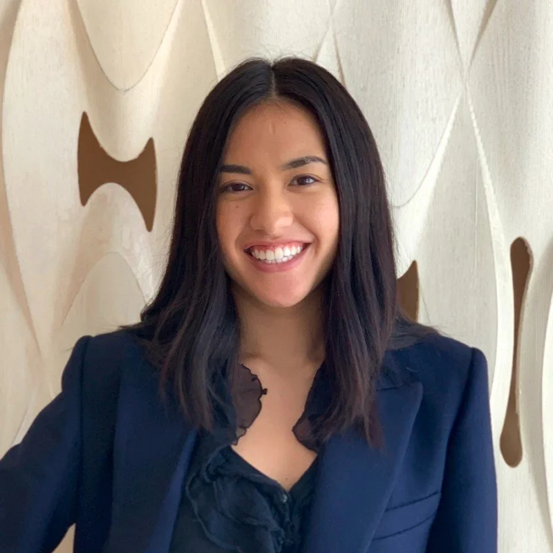 A woman with shoulder-length dark hair, wearing a dark blazer and black ruffled blouse, smiling in front of a beige wall with abstract cutouts.
