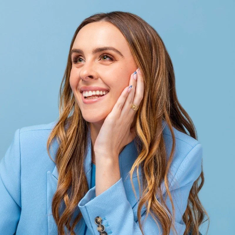 A young woman with long, wavy brown hair, smiling and touching her face with her left hand, wearing a light blue blazer against a blue background.