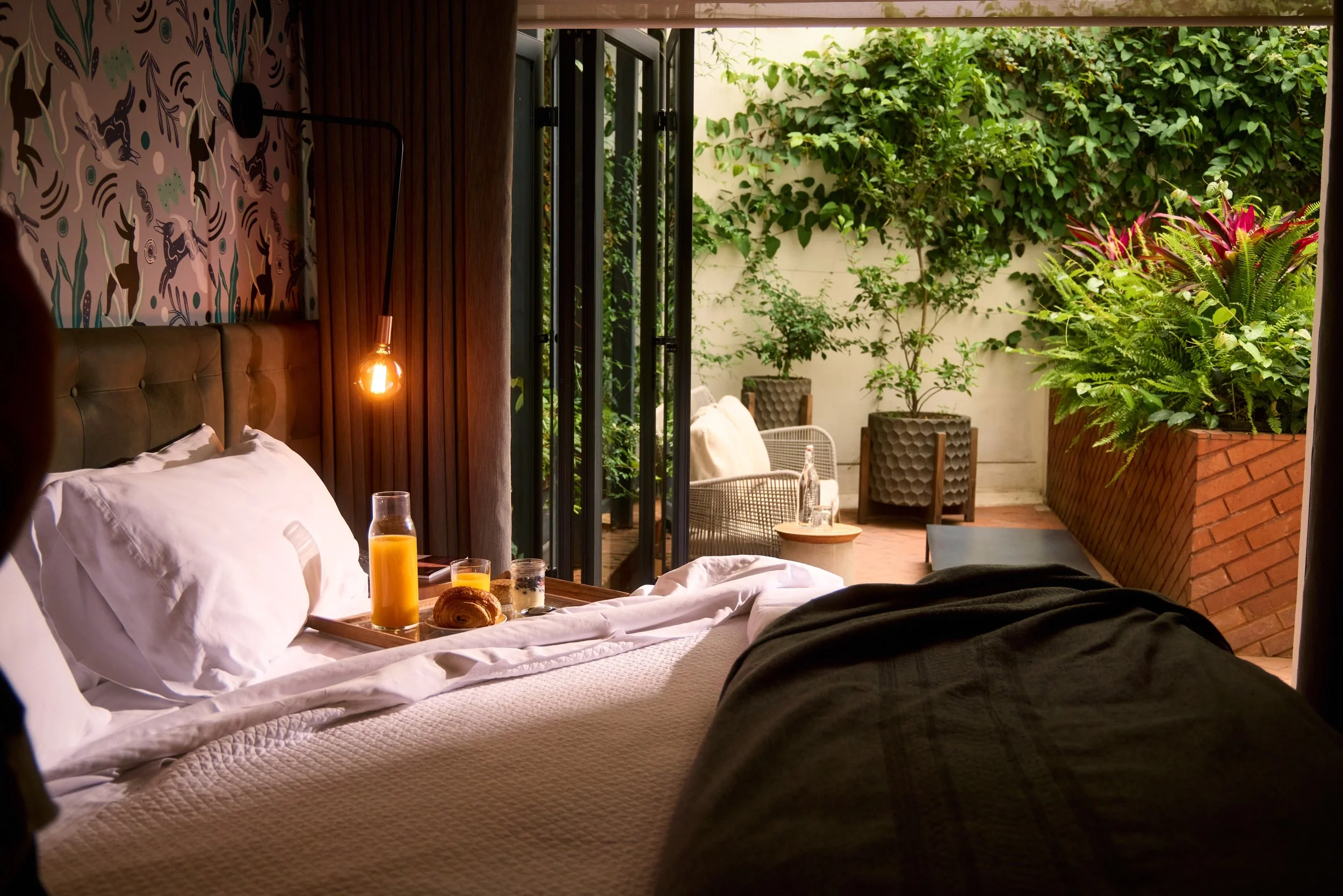 Guest room with breakfast in bed, floor-to-ceiling doors opening to a private garden terrace, and lush greenery.