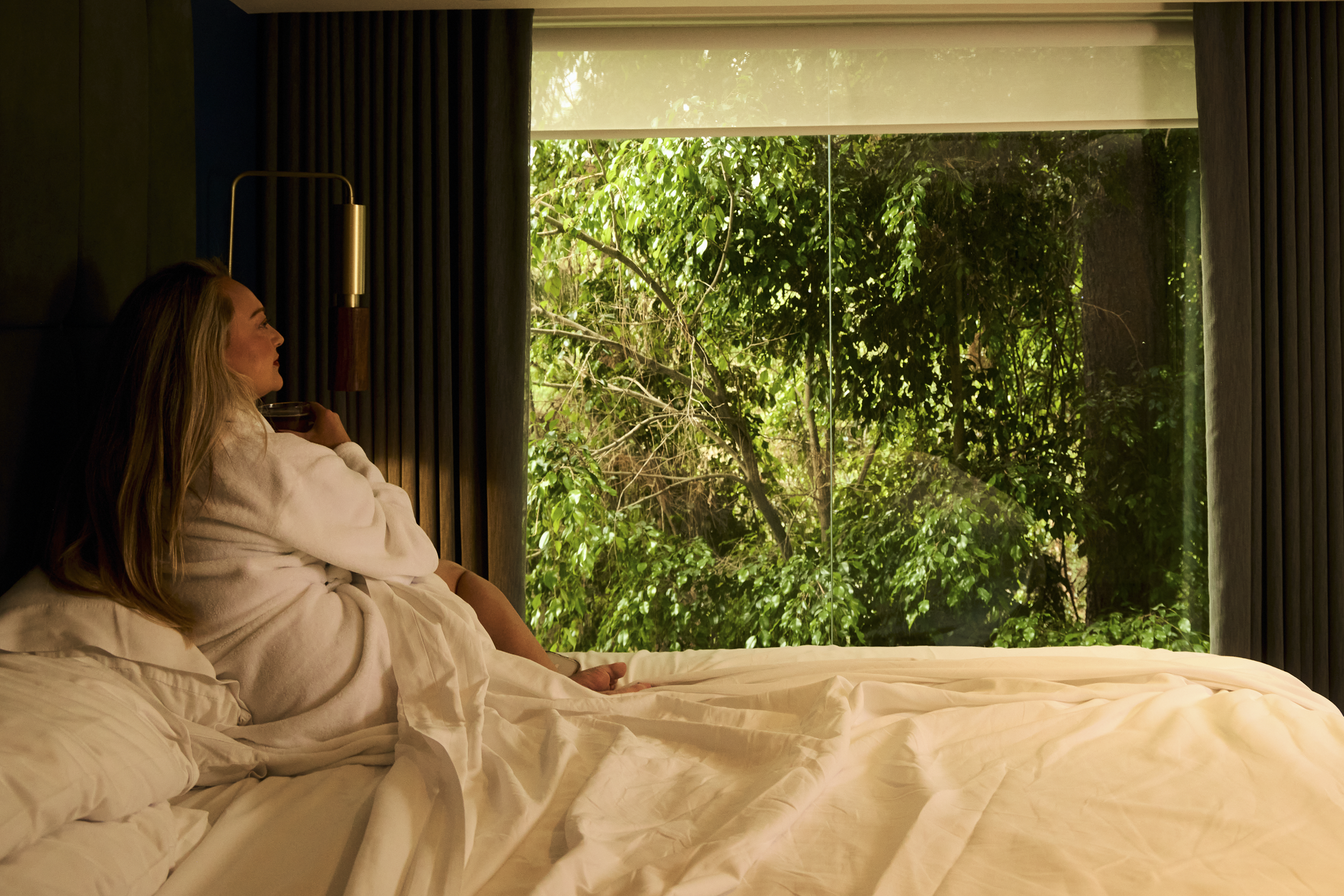 Guest relaxing on a bed in a luxury Campos Residence suite overlooking a private garden through floor-to-ceiling windows.