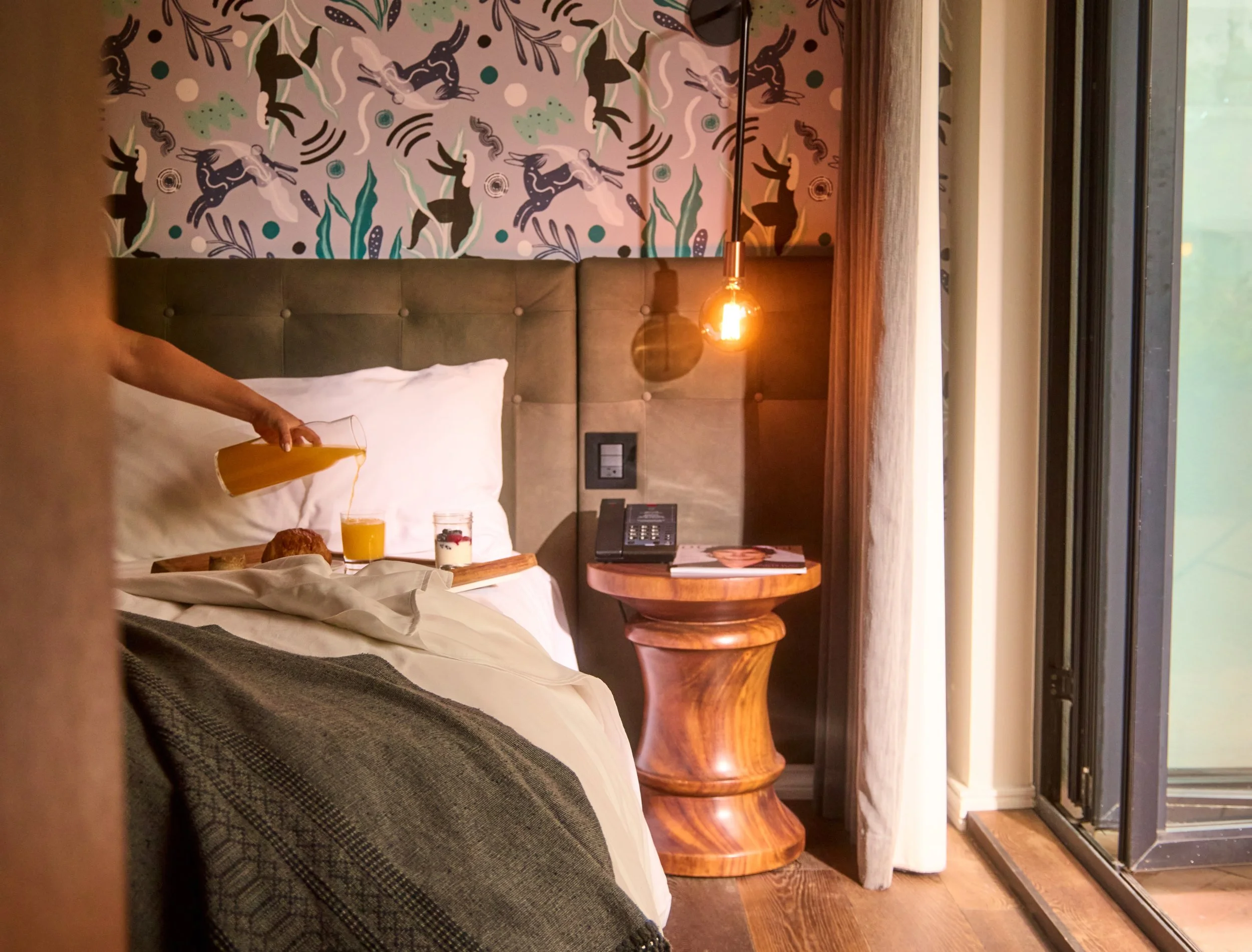 A hotel room with a bed, a nightstand, and a window with curtains. The back wall has patterned wallpaper featuring abstract animal and nature designs in black, white, green, and purple. There is a hanging lamp with an exposed bulb above the nightstan