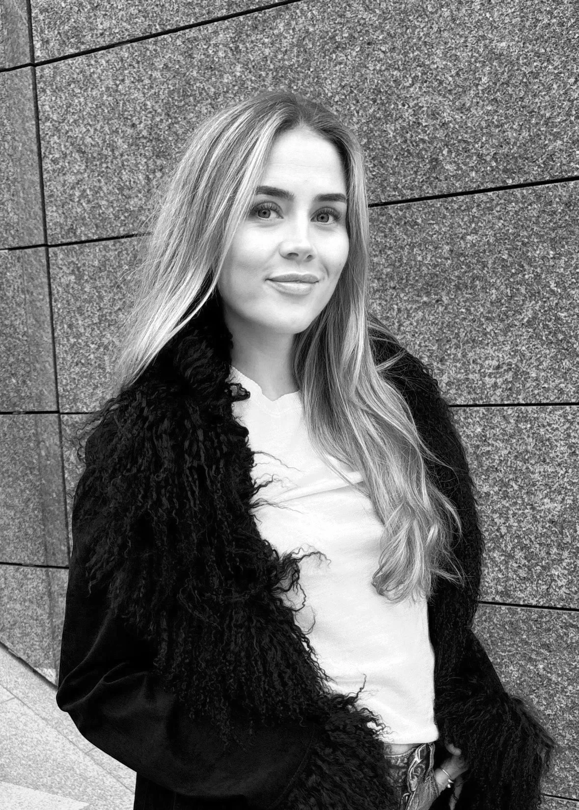 Black and white photo of a young woman with long, wavy hair, standing against a textured wall, wearing a white top and a dark, textured coat.