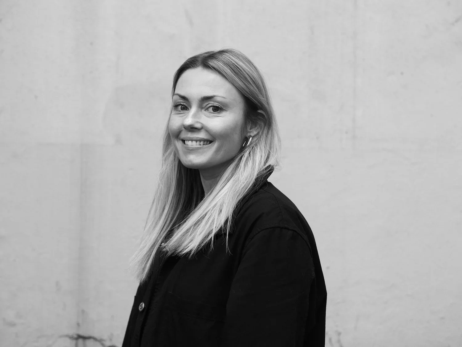 Black and white portrait of a smiling woman with long hair, wearing a dark jacket, standing against a plain wall.