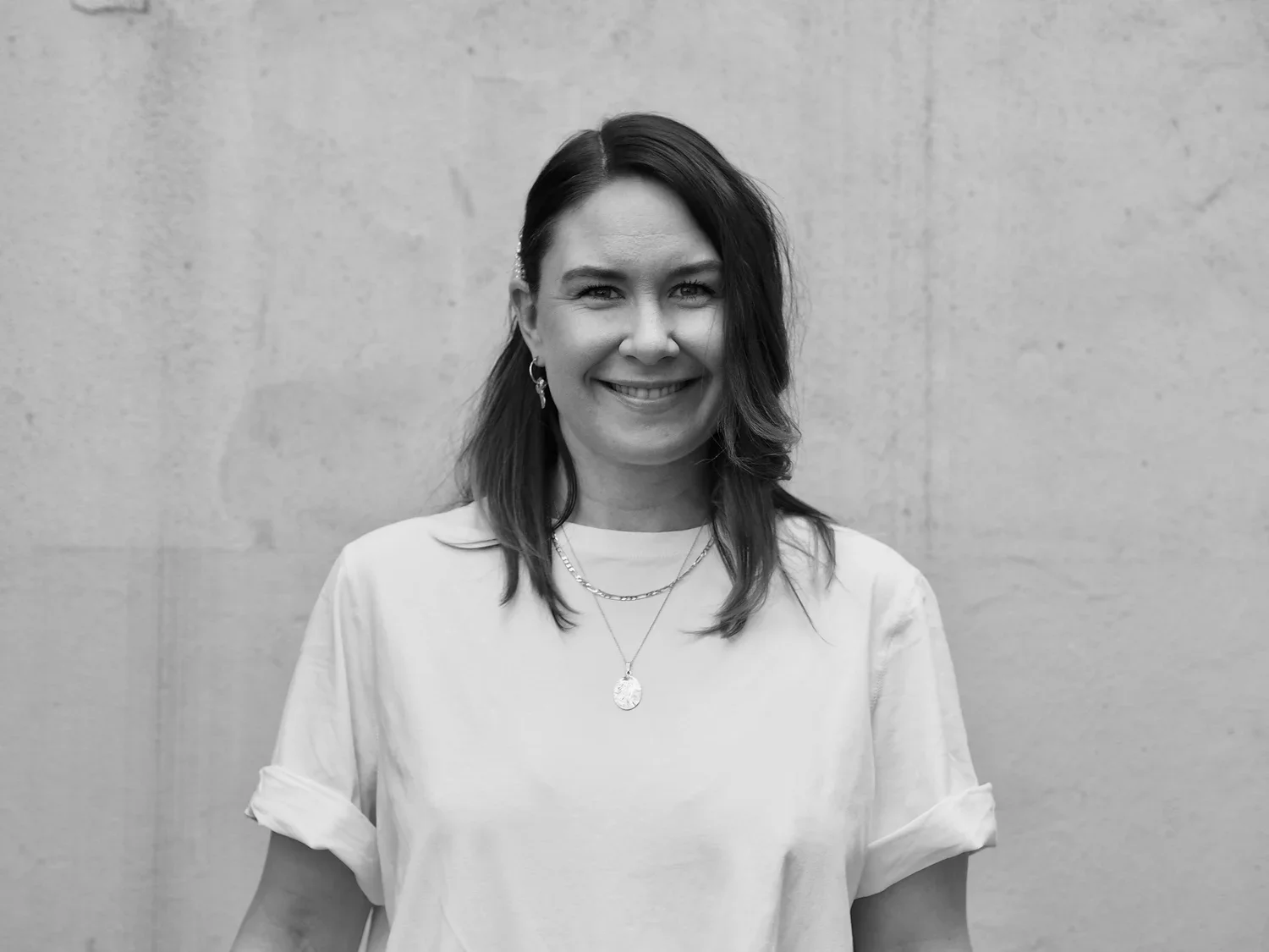 Black and white photo of a smiling woman with shoulder-length dark hair, wearing a white top, layered necklaces, and earrings, standing in front of a plain wall.