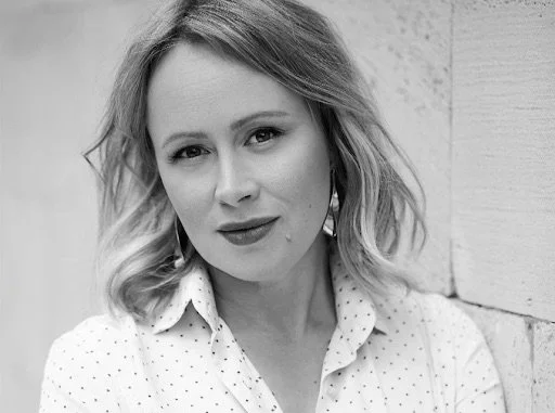 Black and white portrait of a woman with shoulder-length wavy hair, wearing a collared shirt with small polka dots, standing near a brick wall.