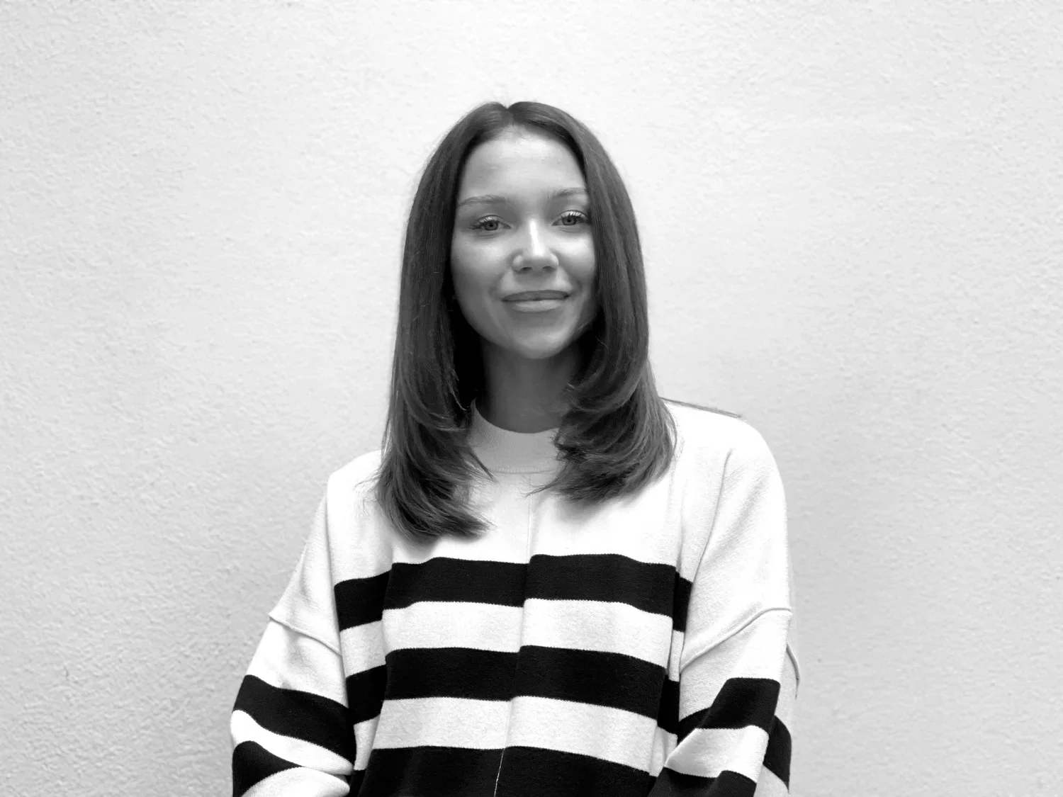 Black and white photo of a young woman with shoulder-length hair, smiling, wearing a striped sweater, standing against a plain textured wall.