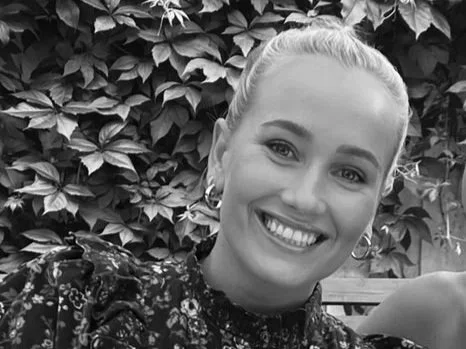 A smiling woman with blonde hair tied back, wearing earrings and a floral top, standing in front of leafy bushes.