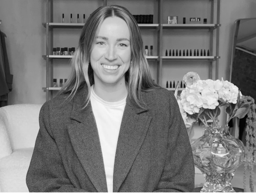 A woman with long hair smiling, wearing a blazer, sitting in a room with a shelf of nail polish bottles and a bouquet of flowers in a glass vase.