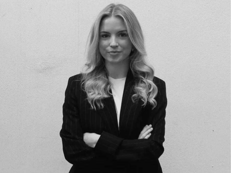 Black and white photo of a young woman with long, wavy hair, wearing a pinstripe blazer and white top, standing against a plain wall with arms crossed.