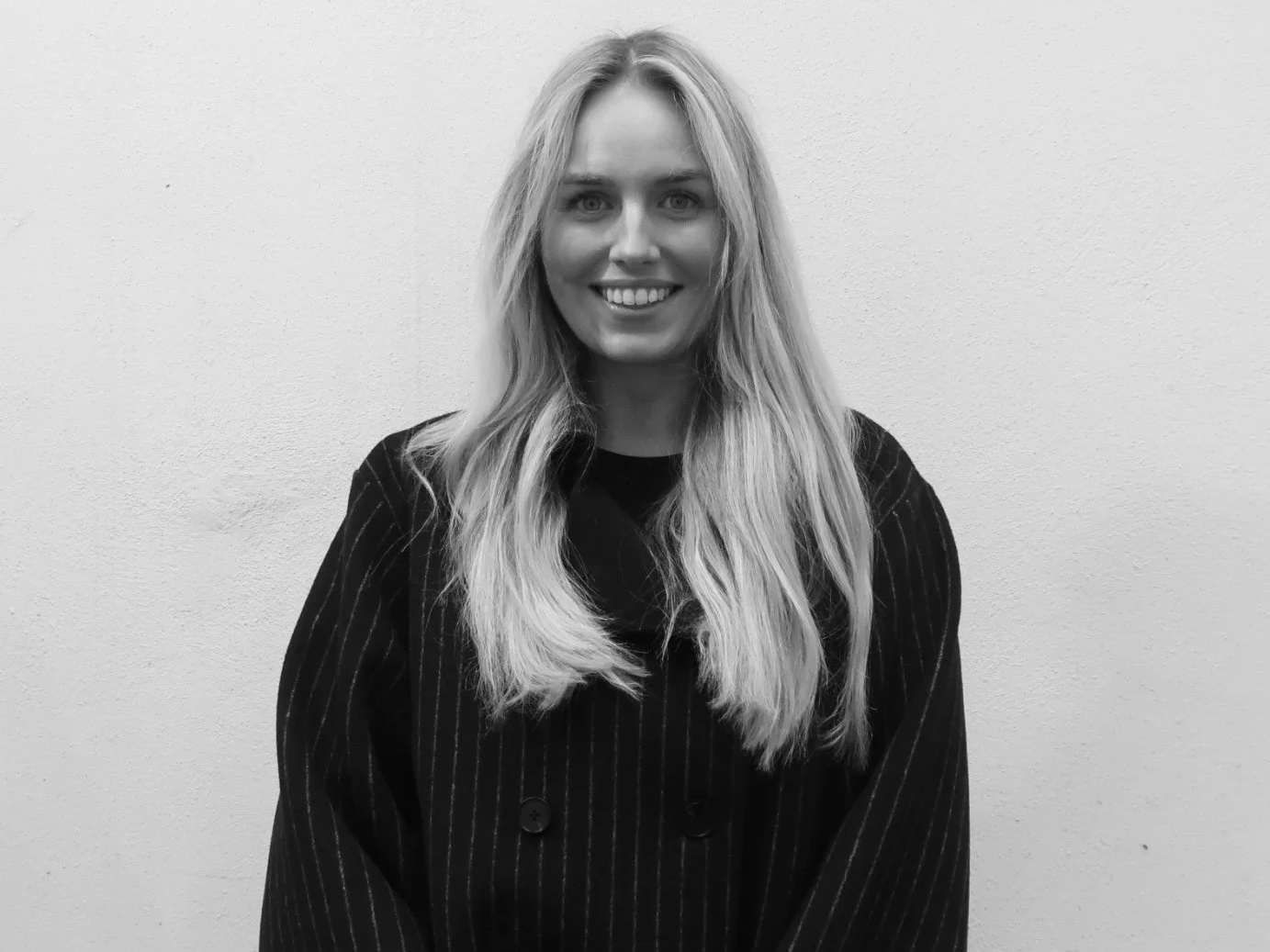 Black and white photo of a smiling woman with long, wavy hair, wearing a dark striped blazer, standing against a plain wall.