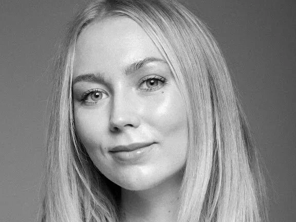 Black and white portrait of a young woman with long, straight blonde hair, light skin, and subtle makeup, smiling softly at the camera.