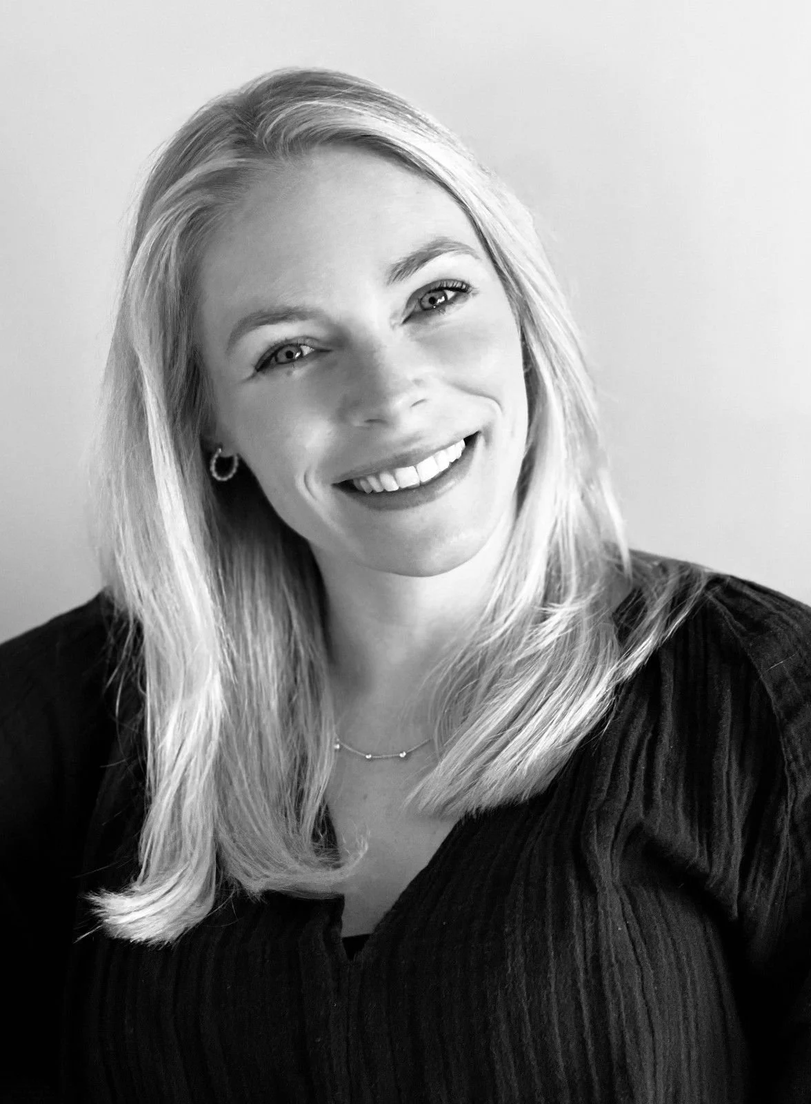A smiling woman with shoulder-length blonde hair, wearing a black top and a delicate necklace, against a plain background.