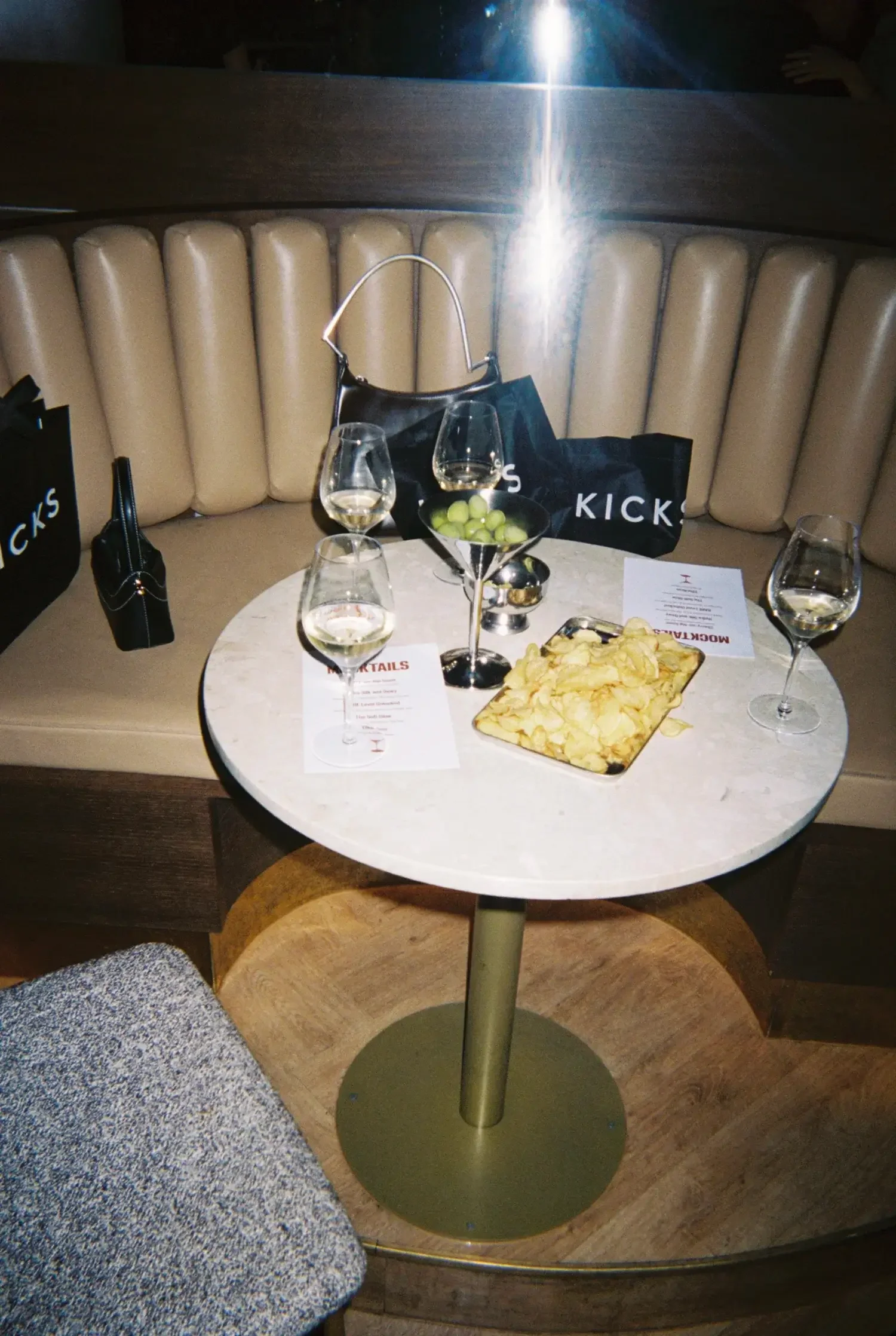 A table with four glasses of white wine, a small bowl of green grapes, a tray of potato chips, and printed menus. The table is in a booth with beige cushioned seating and black shopping bags in the background.