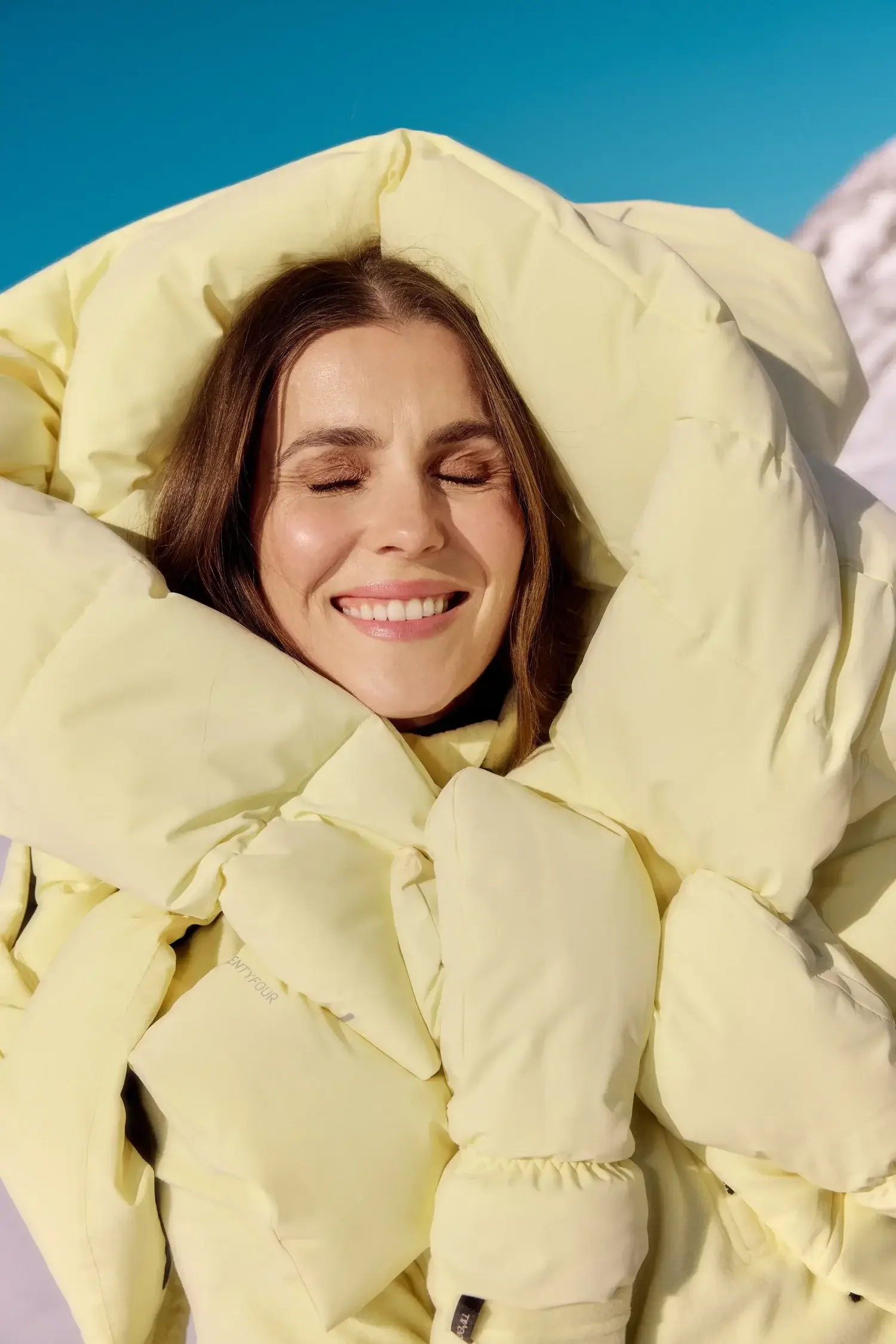 A woman with brown hair and closed eyes smiling, bundled in a light yellow puffy jacket, with snow and a blue sky in the background.