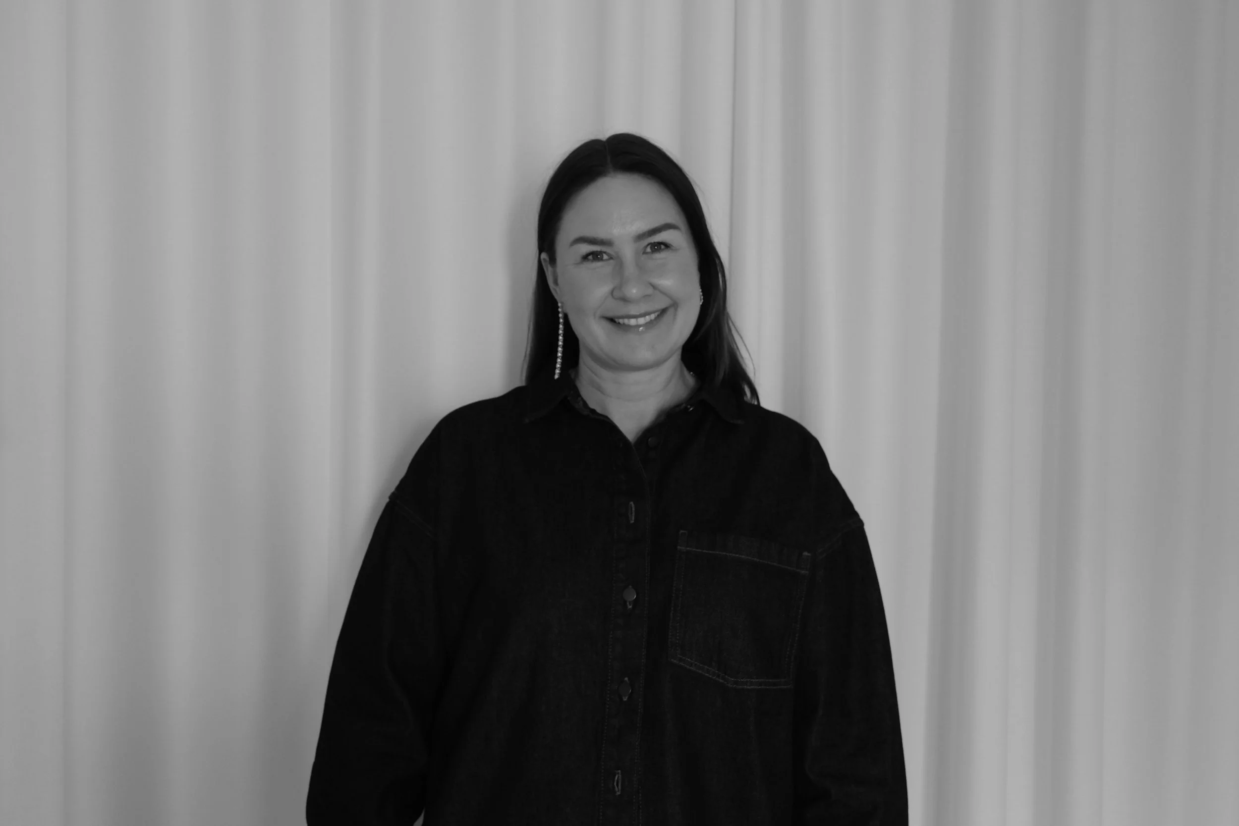 Black and white photo of a smiling woman with shoulder-length dark hair, wearing a white top, layered necklaces, and earrings, standing in front of a plain wall.