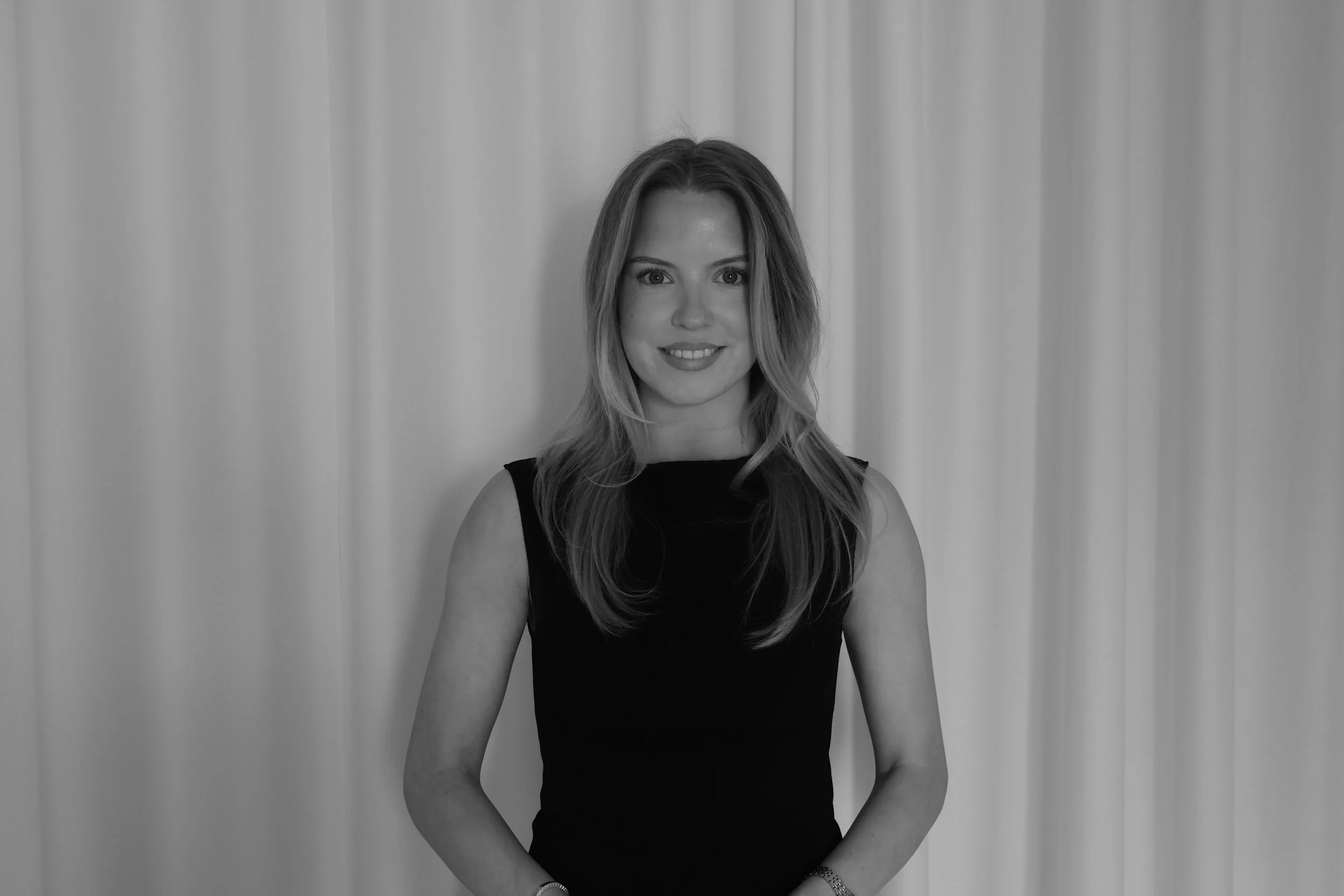 Black and white photo of a young woman with long, wavy hair, wearing a pinstripe blazer and white top, standing against a plain wall with arms crossed.