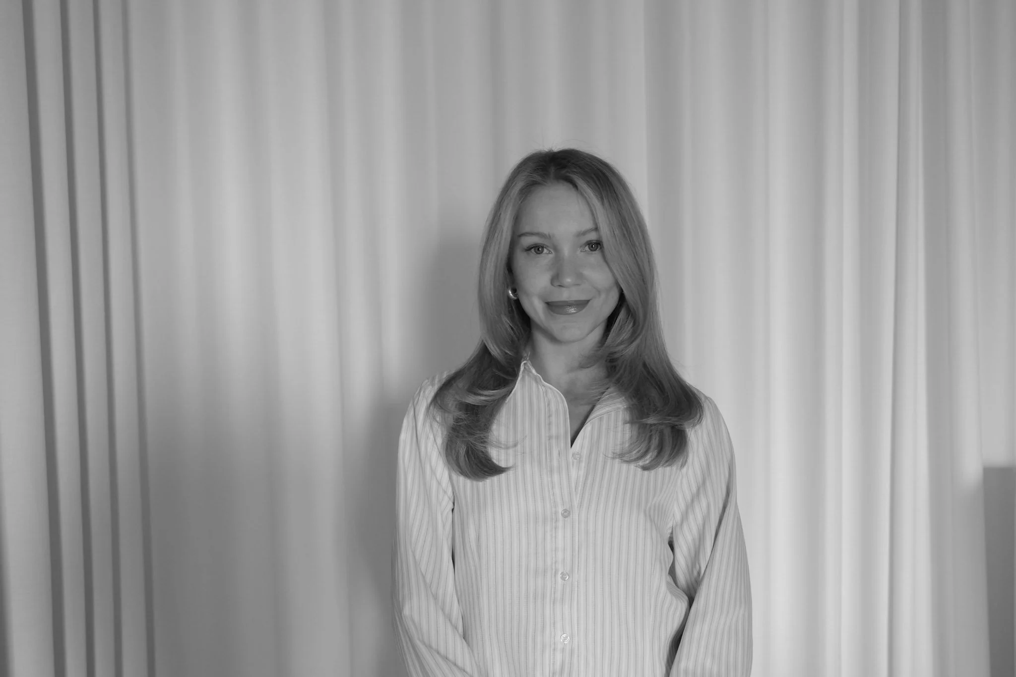 Black and white photo of a young woman with shoulder-length hair, smiling, wearing a striped sweater, standing against a plain textured wall.