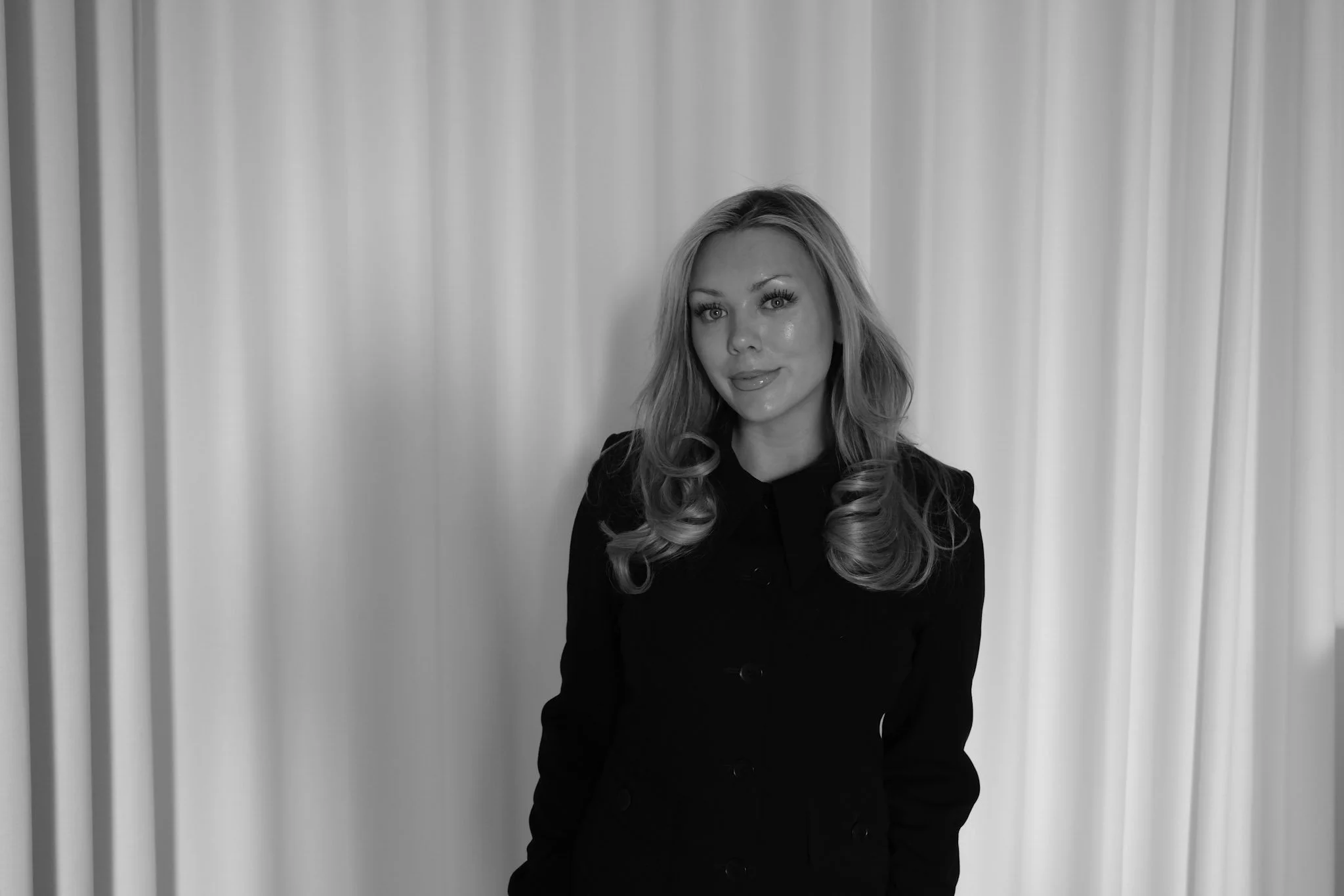 Black and white photo of a woman with blonde hair, wearing a blazer and standing against a plain wall.