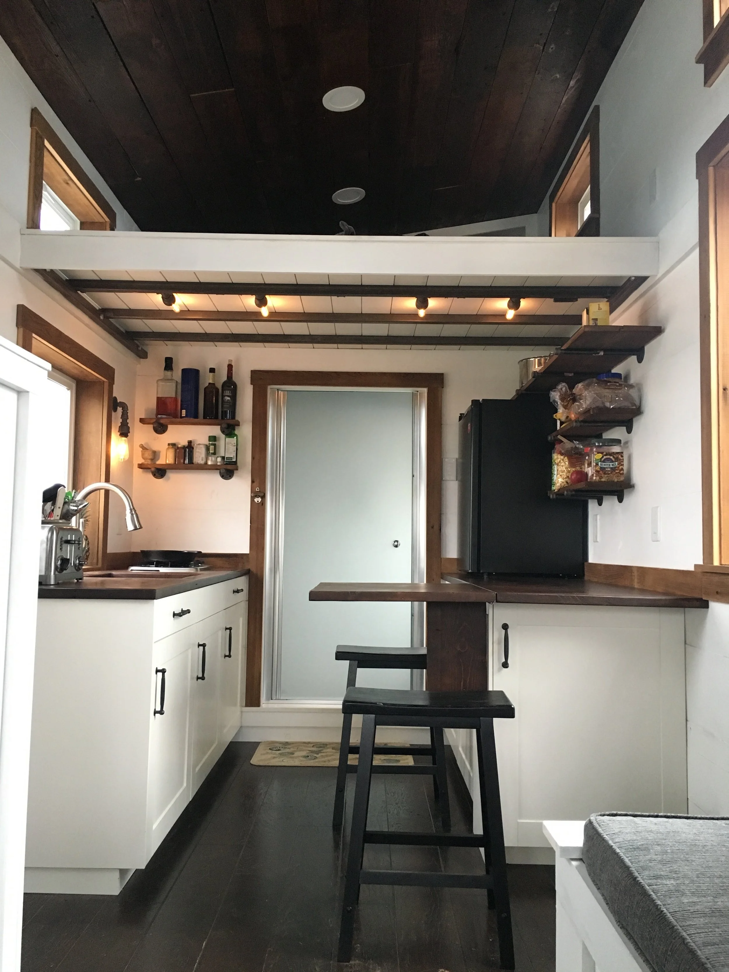 Interior view of a custom mobile tiny home by Prenvalley Builders featuring a compact kitchen, built-in dining surface, open shelving, and a lofted sleeping area designed to maximize vertical space.