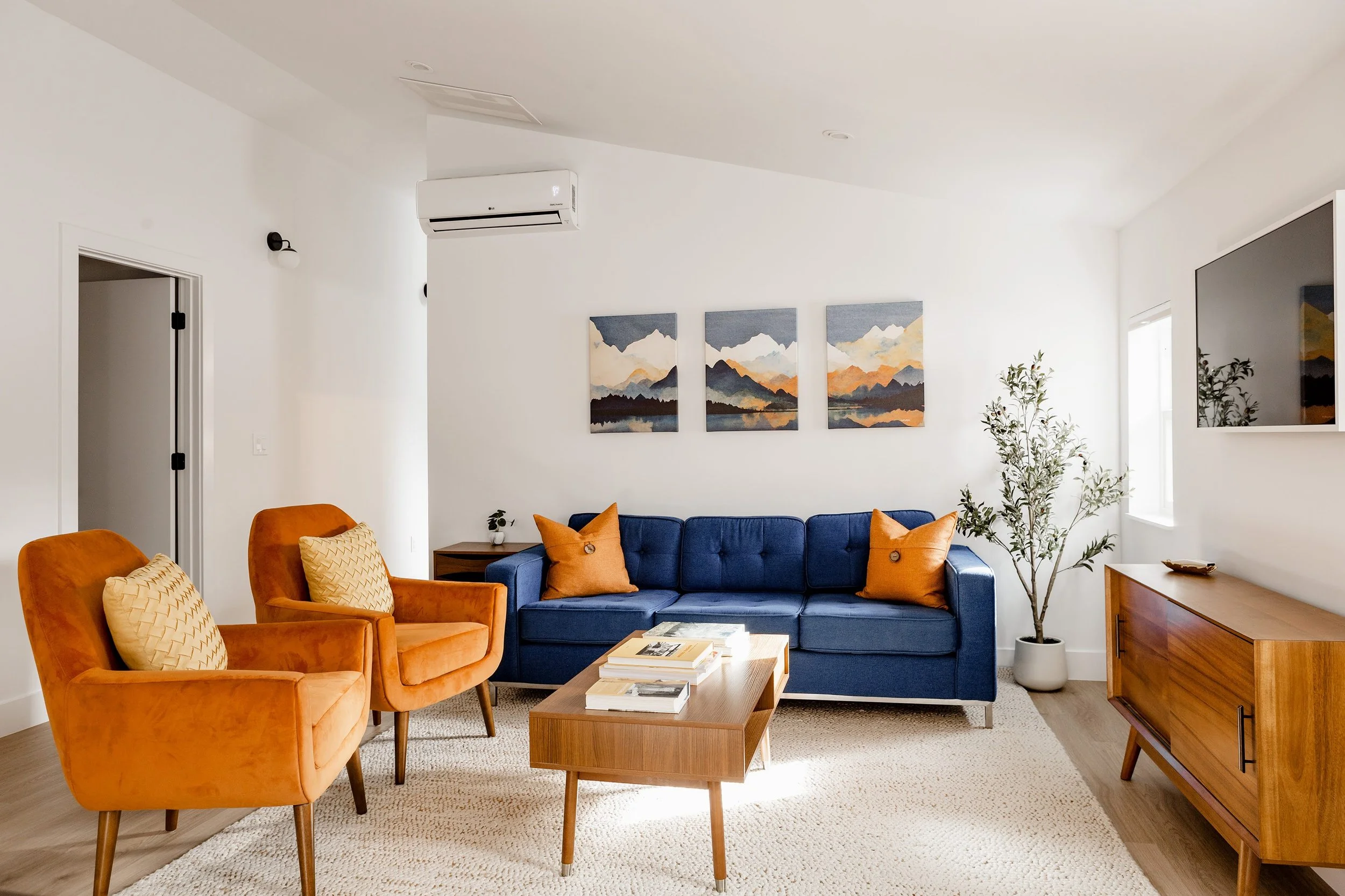 Wide view of a modern Denver ADU living room featuring a blue sofa, orange accent chairs, wood coffee table, and curated artwork by Prenvalley Builders.