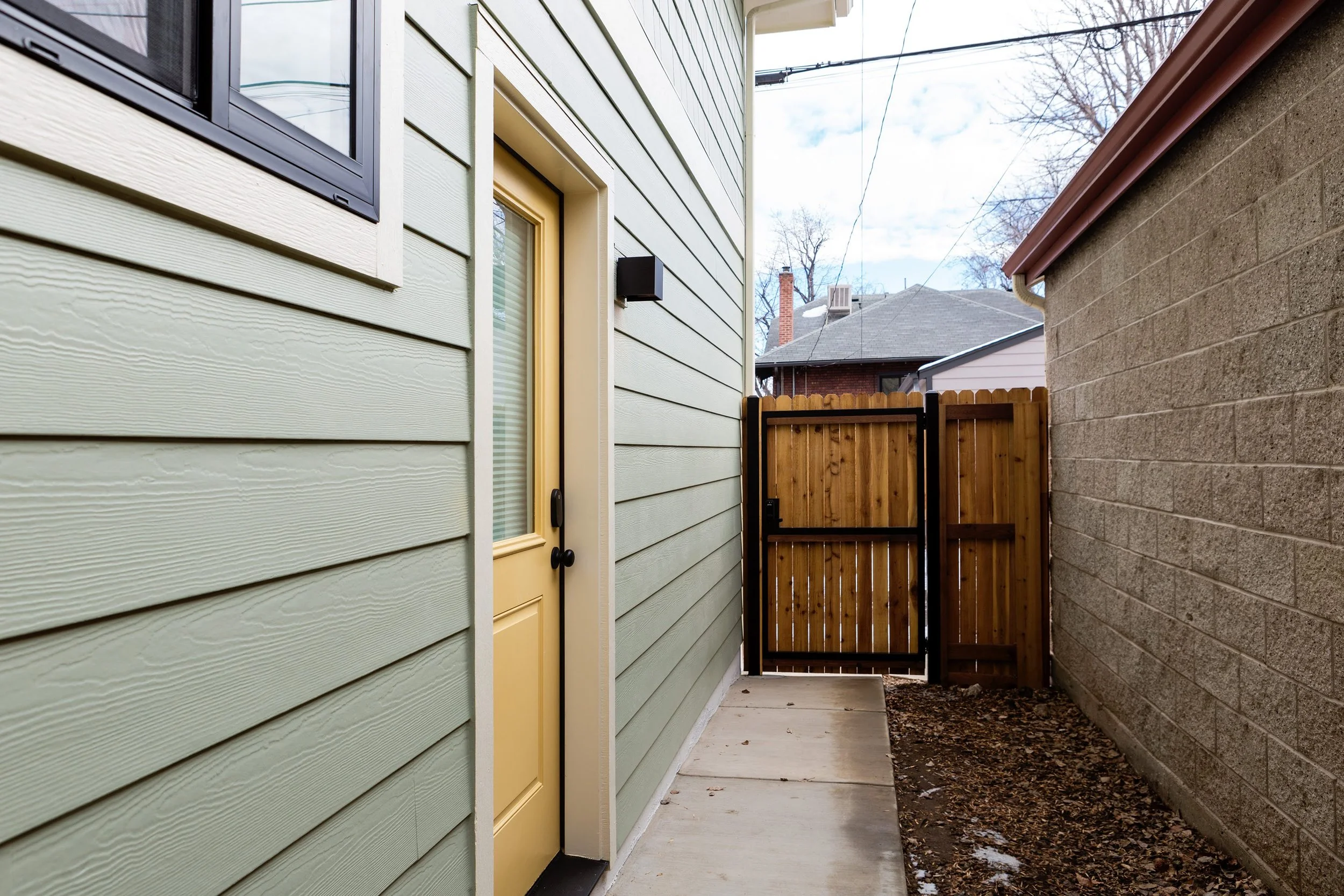 Denver ADU exterior side entry with a private door, horizontal siding, concrete walkway, and wood privacy gate.