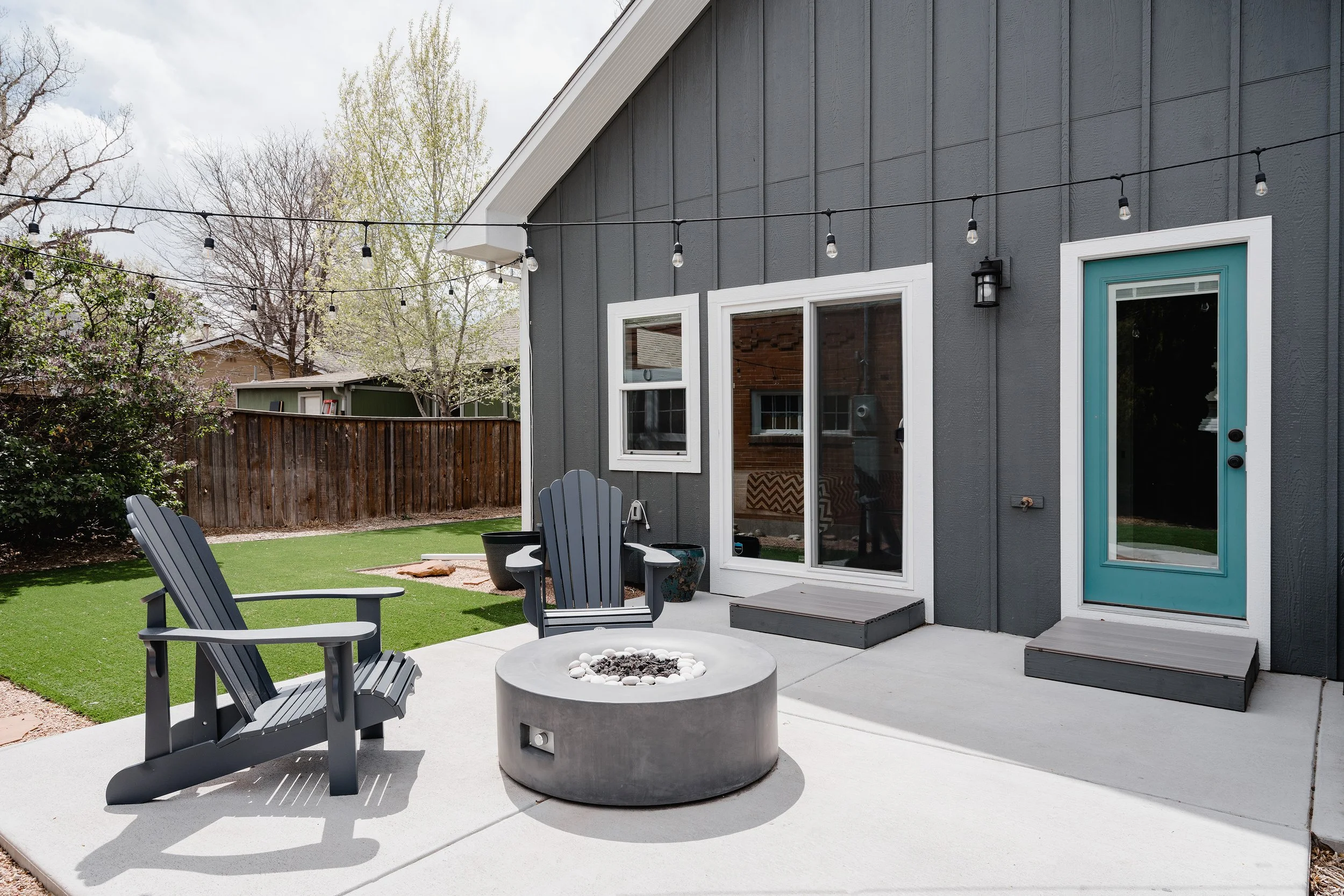 Exterior view of a Denver primary-suite addition showing board-and-batten siding, sliding glass doors, a private concrete patio, and a built-in gas fire pit.