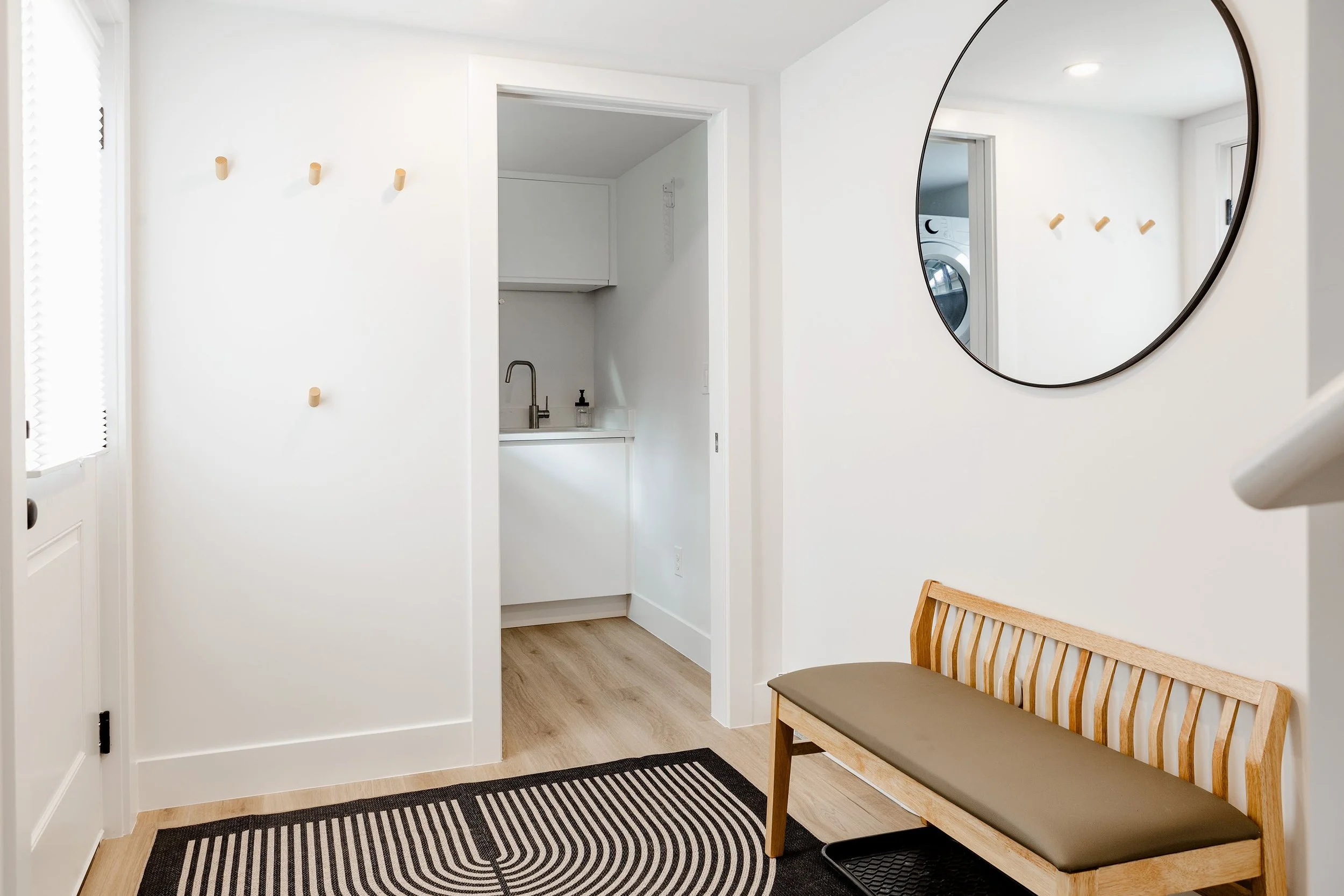 Bright Denver ADU entryway featuring a built-in bench, wall hooks, round mirror, and a compact laundry nook with integrated sink.