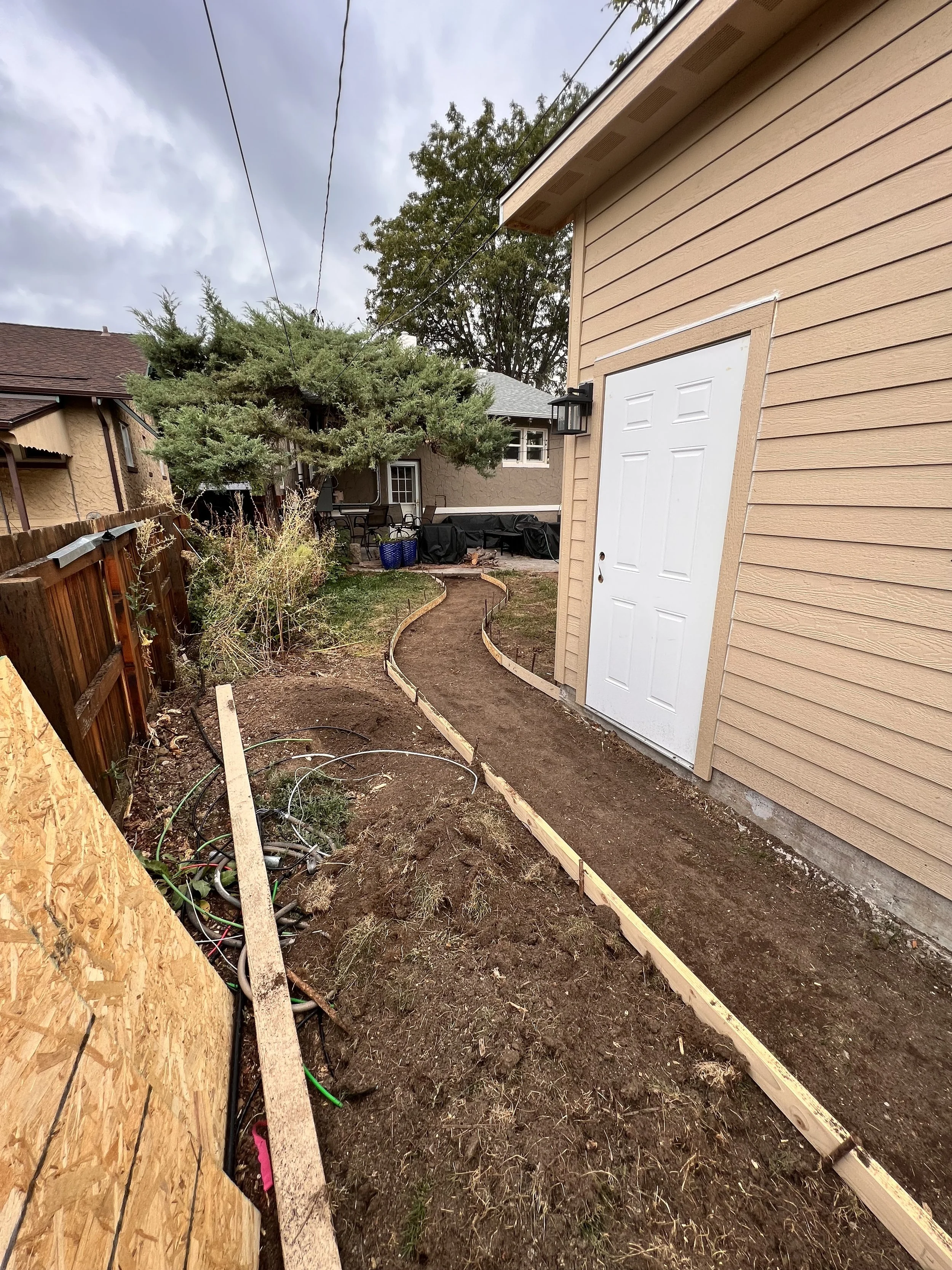 Side yard site work in progress for a new custom garage in Denver, Colorado, showing graded pathways, formwork for a concrete walkway, and exterior access planning by Prenvalley Builders.