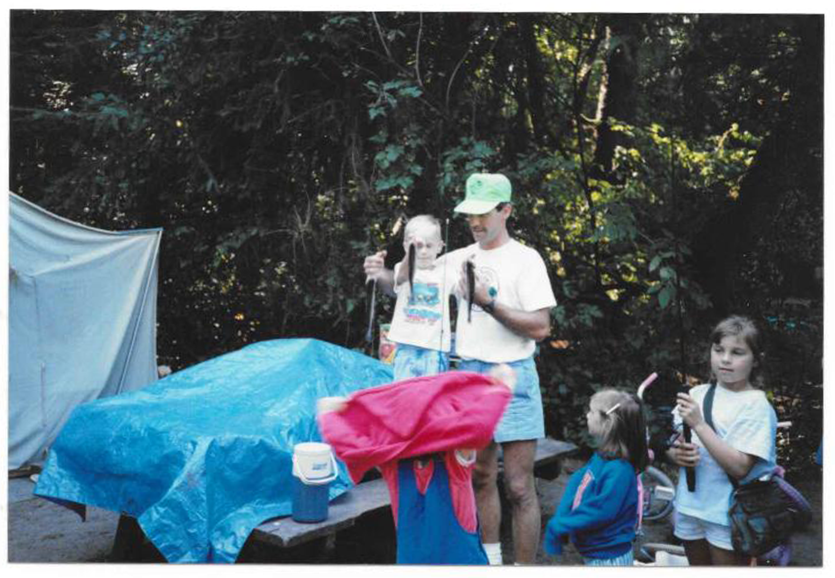 A group of children and an adult camping outdoors near a wooded area. The children are holding small fish, with a makeshift tent and camping supplies visible.