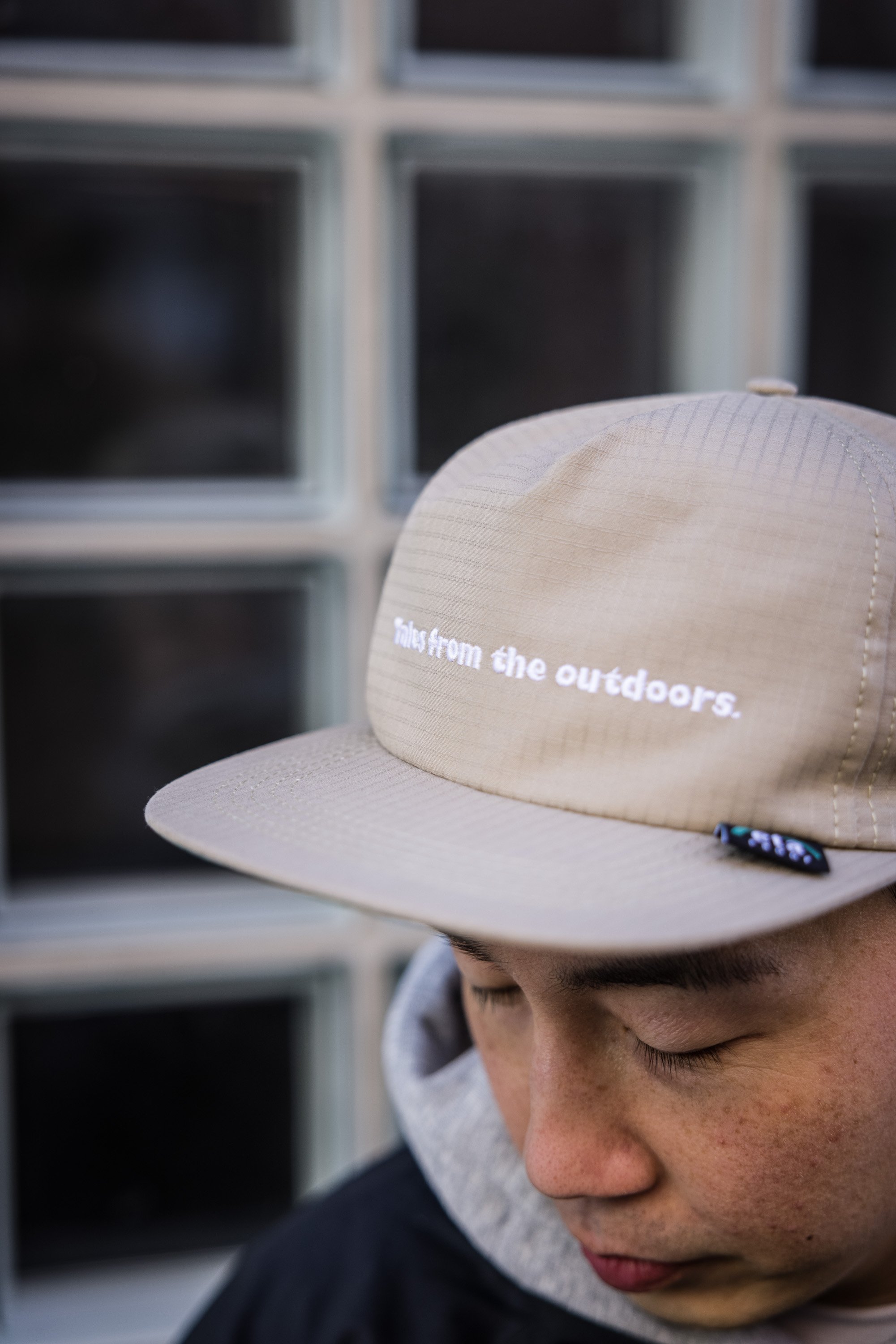 Close-up of a man wearing a beige cap with the text 'tabs from the outdoors' in white. The man's face is partially visible with closed eyes and freckles, and he is wearing a gray hoodie and black jacket. The background shows a window with multiple glass blocks.