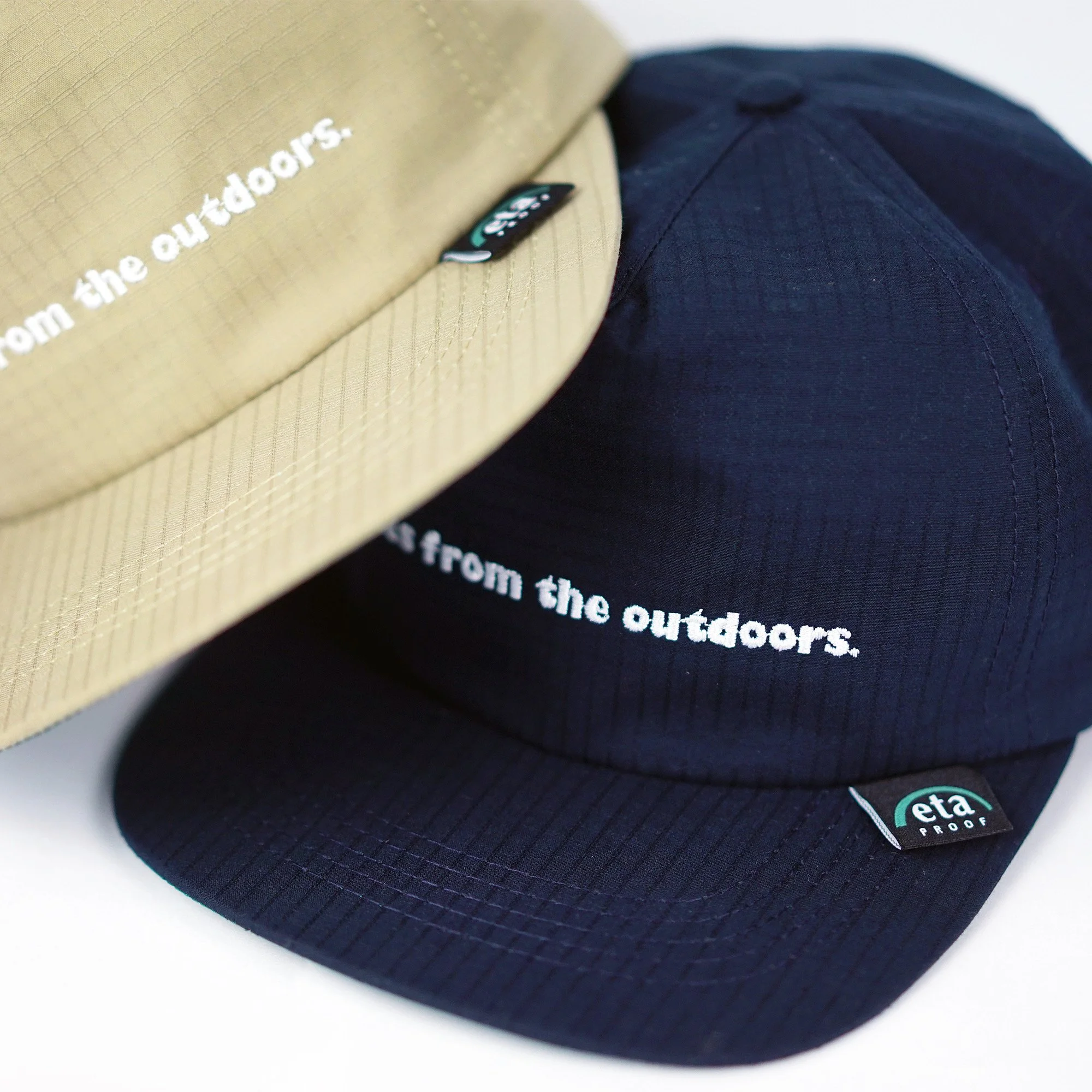 Close-up of two outdoor caps, one beige and one black, with the phrase 'from the outdoors.' embroidered on the front.