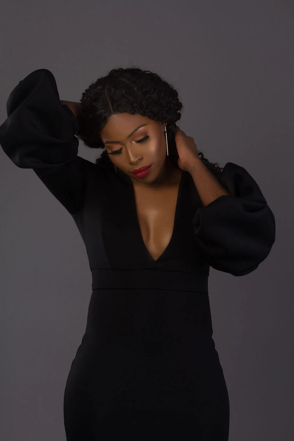 Woman in black dress with voluminous sleeves, striking a pose with hands on head, against gray background.