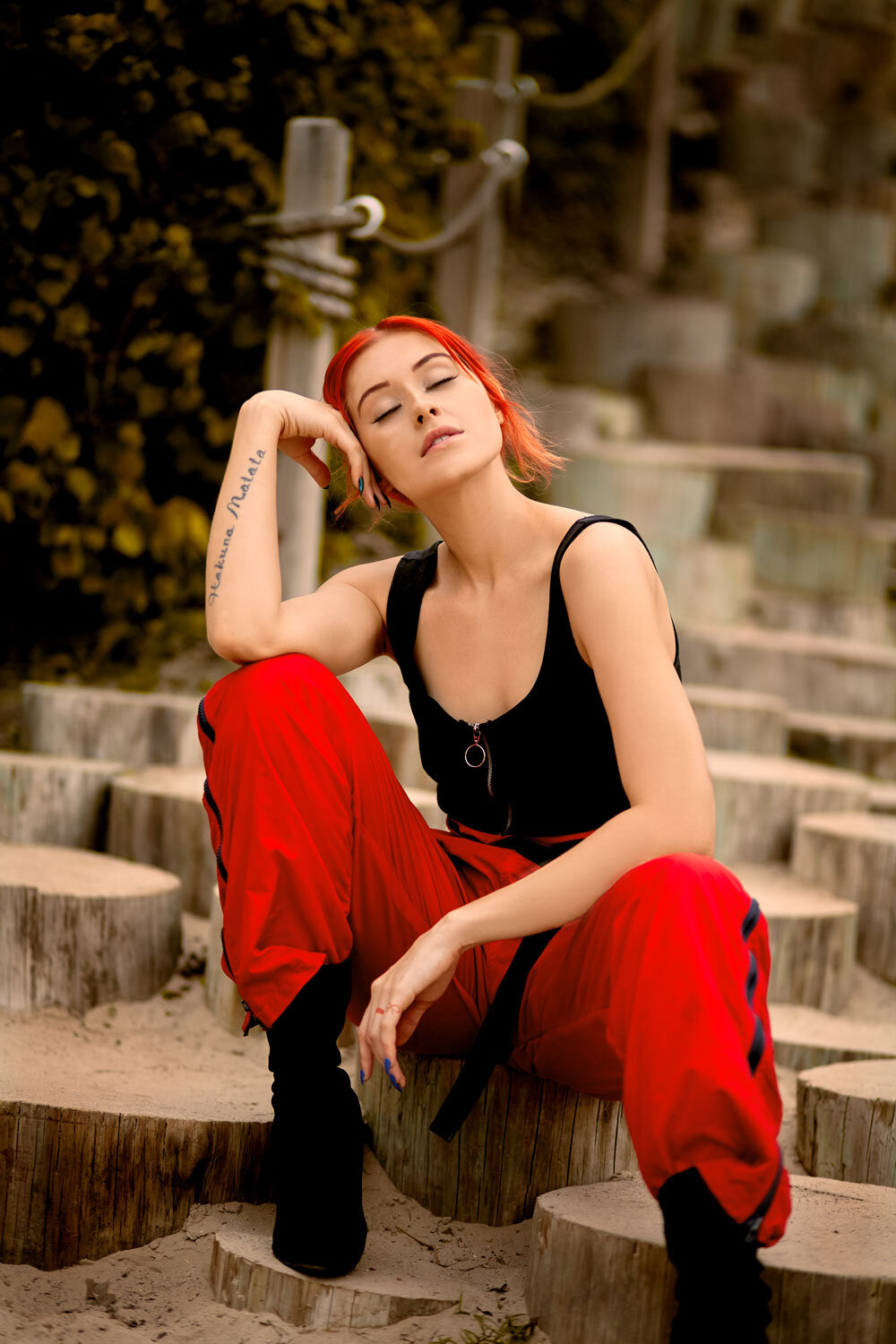 A person with red hair is sitting outdoors on wooden steps, wearing a black top and red pants. They have one knee up and are resting their elbow on it, with eyes closed and a relaxed expression.