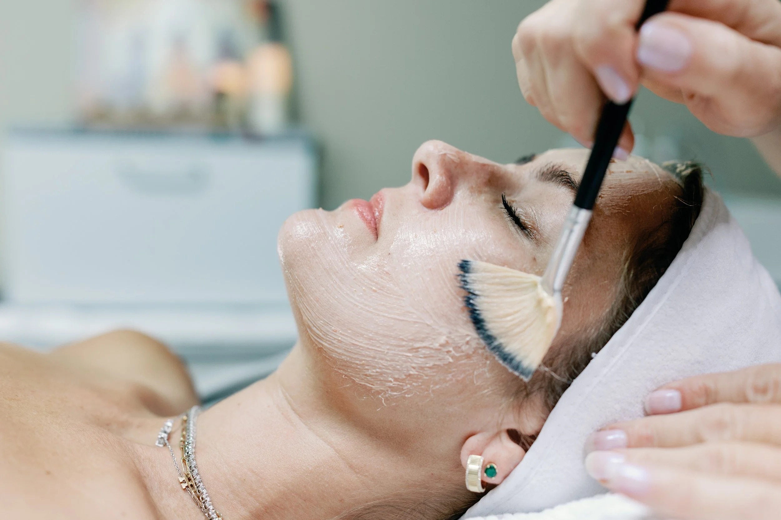 Woman receiving a facial masque applied with a fan brush in a spa setting, wearing a headband.