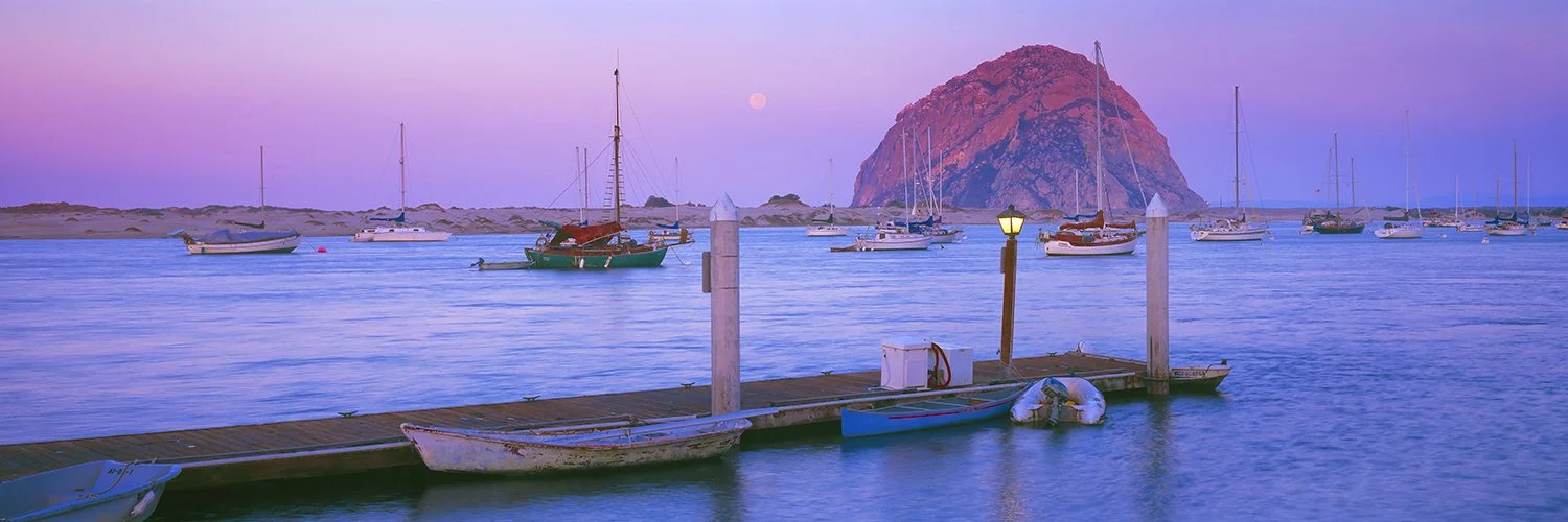 Morro Bay harbor at dusk with sailboats, docks, Morro Rock and the moon.