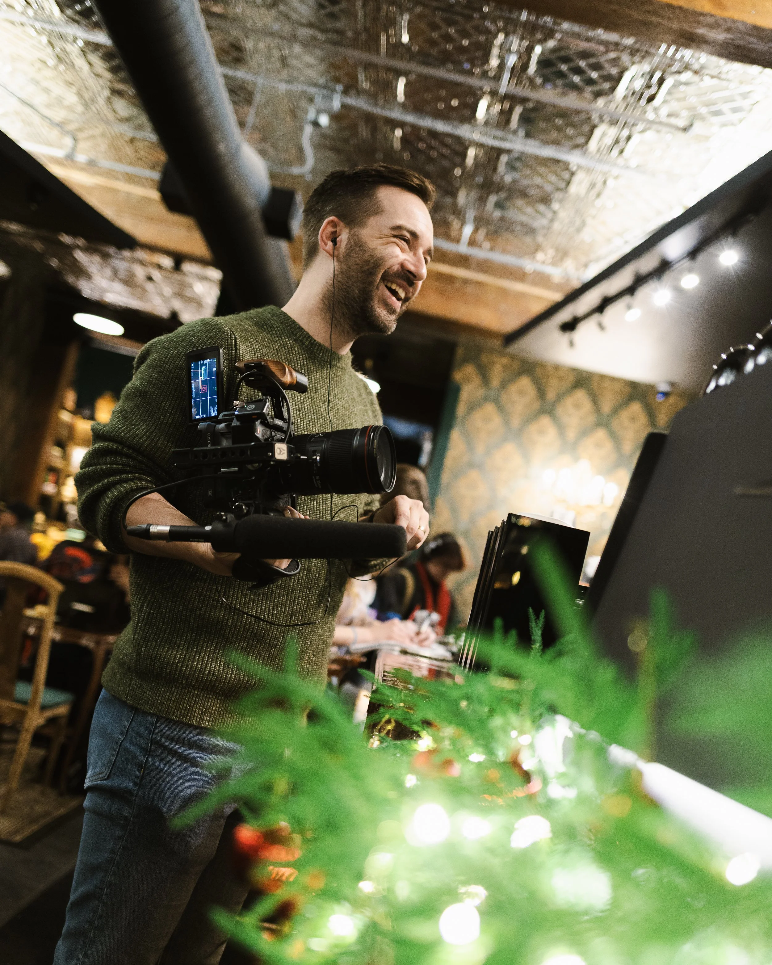 A man with short dark hair and a beard, wearing a green sweater and jeans, smiling and laughing while looking at a computer screen. He has earphones in his ears and is holding a camera with a monitor attached. The background shows an indoor setting with warm lighting and wooden ceiling, and some greenery in the foreground.
