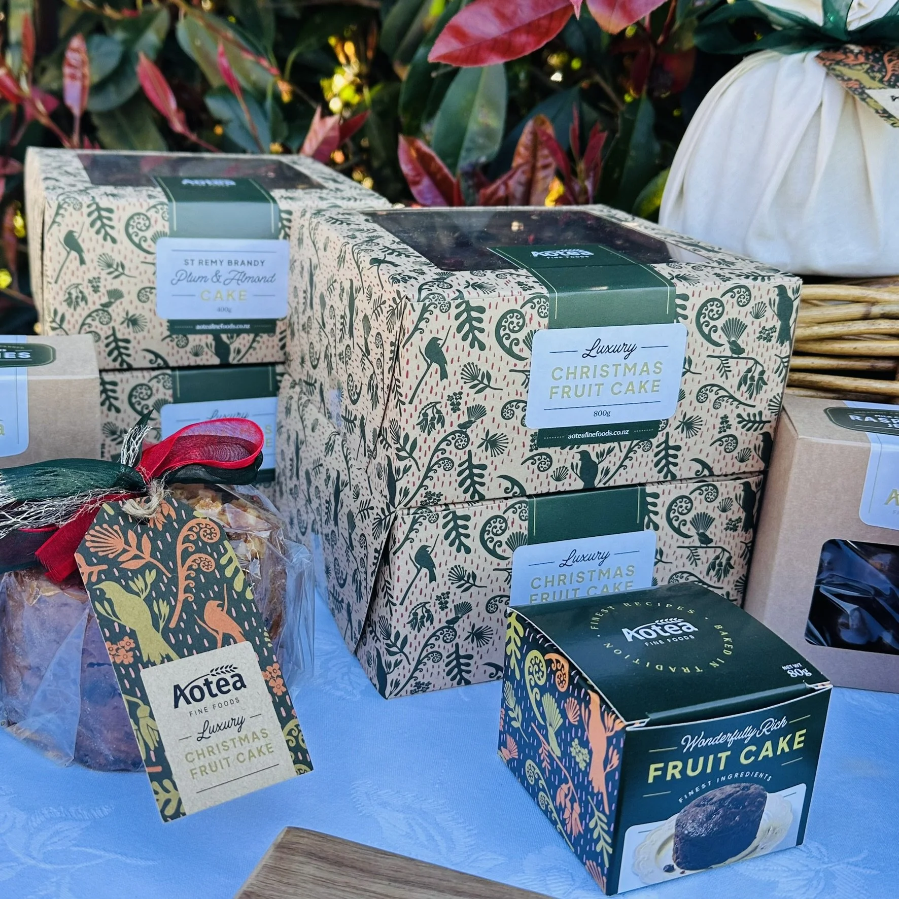 Boxes of luxury Christmas fruit cake from Aotea Fine Foods on a table with greenery in the background.
